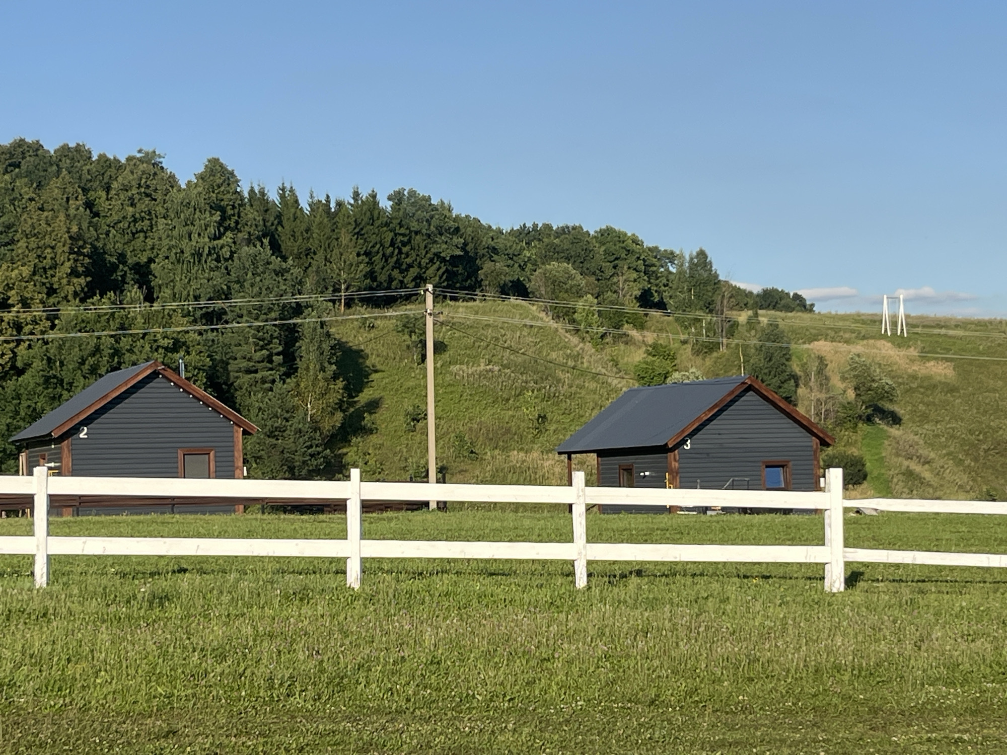 A serene rural landscape under a clear blue sky features two dark gray wooden cabins with dark roofs and brown trim. The cabin on the left is numbered '2', and the cabin on the right is numbered '3'. A white wooden fence, with horizontal planks supported by vertical posts, spans the foreground of the image, separating a vibrant green grassy field from the area where the cabins are situated. Beyond the cabins, rolling hills are covered in a mix of lush green grass and dense trees. Utility poles and wires are visible on the hillside, and in the far distance, a white structure resembling a swing set or a large archway stands on the crest of a hill. The scene appears to be located in Srednie Pinyachi, Russia, conveying a sense of peaceful countryside living.
