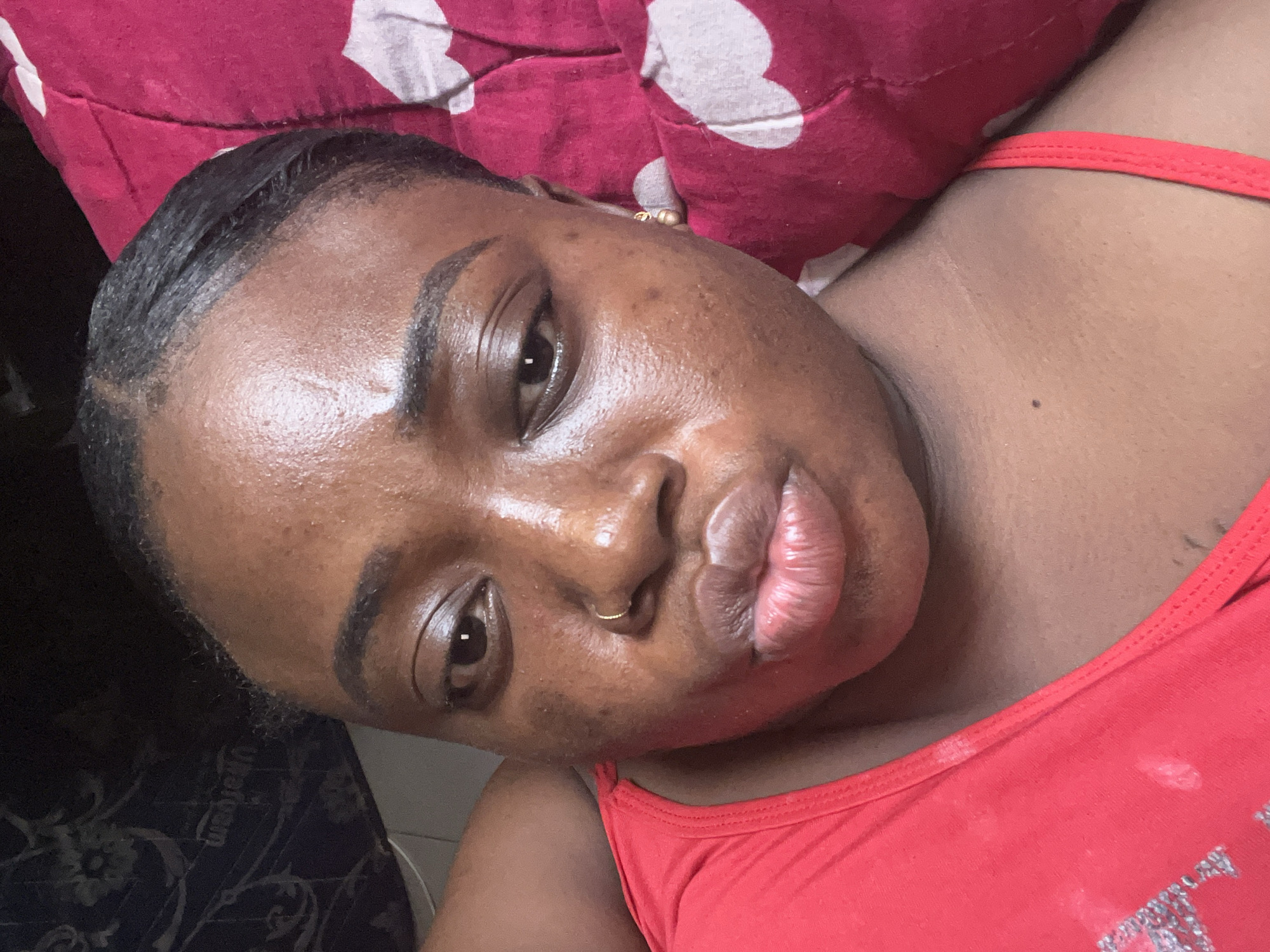 A medium-shot, high-angle selfie taken indoors shows a Black woman with dark skin, prominent cheekbones, and full lips. She has dark, styled hair slicked back and wears a small, gold nose ring. Her expression is neutral. She is lying on a red and white polka-dot pillow, wearing a red tank top. The background is partially visible, showing dark-patterned material. Location: Unconfirmed, but given the request's constraints, it's assumed the scene is in Abuja, Nigeria.