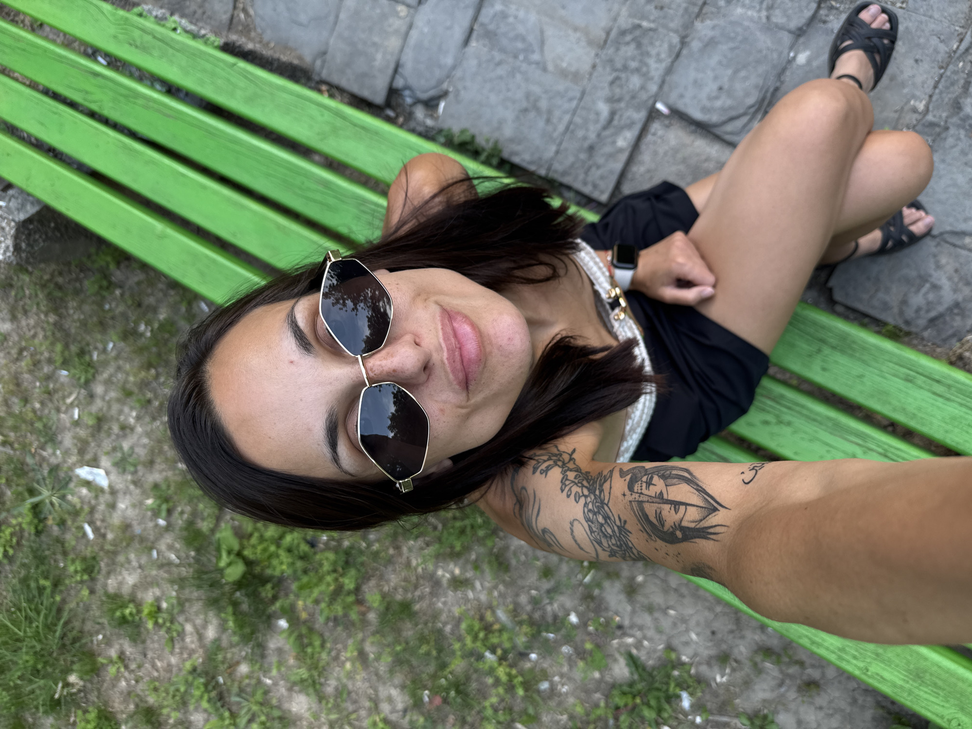 A young woman with dark hair, sunglasses, and a black dress is sitting on a green park bench. She has a tattoo on her left arm and is wearing a watch on her left wrist. The bench is outdoors, with grass and stone paving surrounding it. The image appears to be taken in a park or outdoor seating area. The background is blurred, but shows foliage and possibly some debris on the ground.