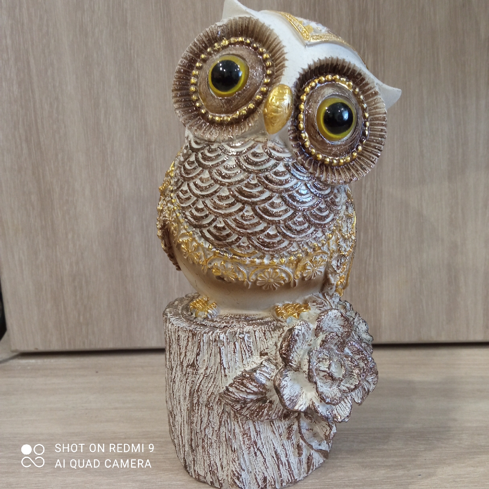 A ceramic owl figurine is positioned on a wooden surface in Chaikovsky, Russia. The owl is white and brown with gold accents, large black eyes, and sits atop a tree stump base adorned with a flower. The background consists of vertical wood paneling. A watermark indicating the image was shot on a Redmi 9 with AI Quad Camera is located in the bottom left corner.