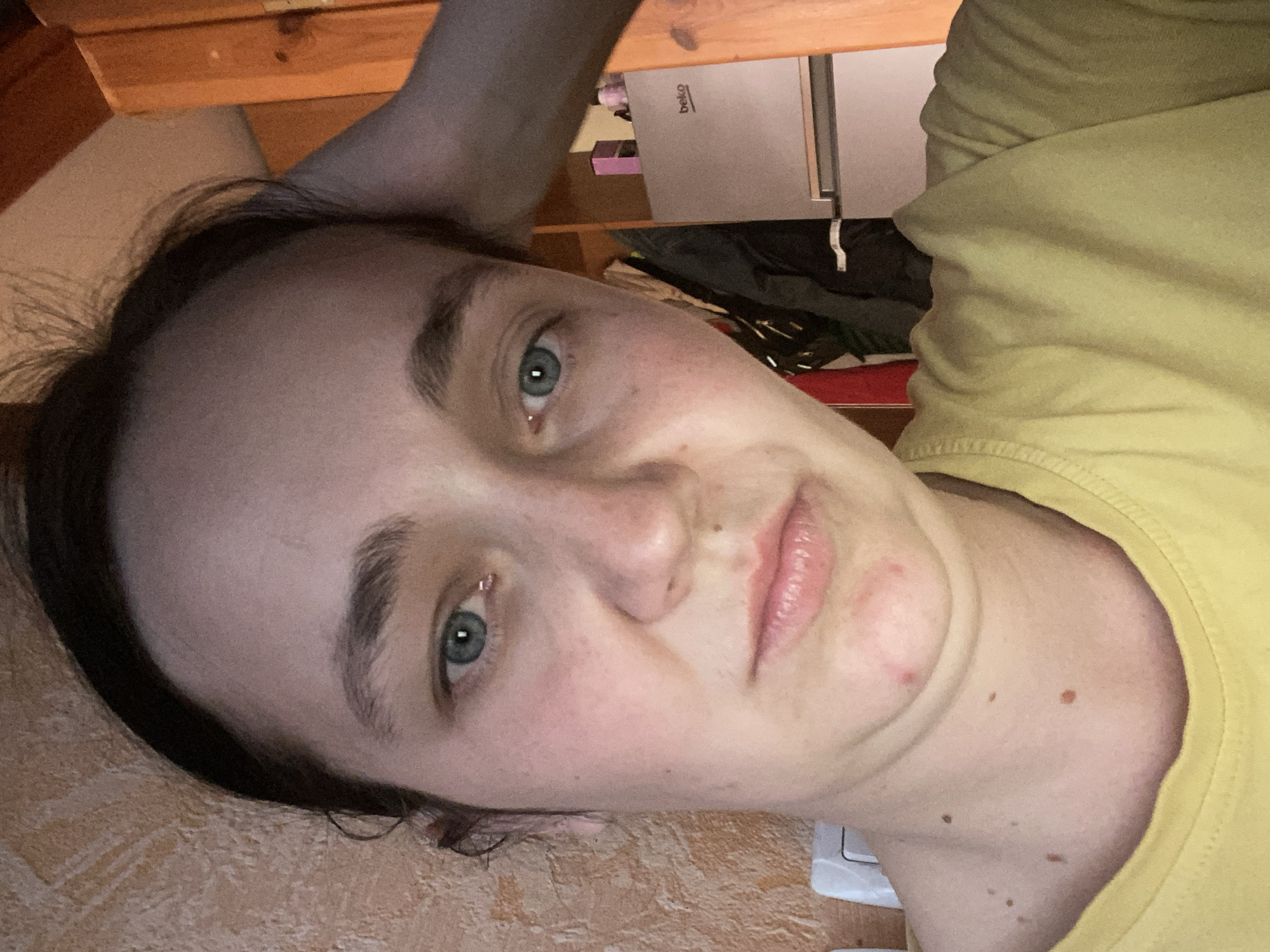 A young woman with dark hair and blue eyes takes a selfie. She is wearing a yellow t-shirt and has her right hand on the back of her head. Her expression is neutral, and she has a few moles on her neck and chin. There is a small red pimple on her chin. In the background, a beige textured wall is visible on the left, and a wooden door frame and a small silver refrigerator are on the right. The lighting suggests an indoor setting, possibly a home.