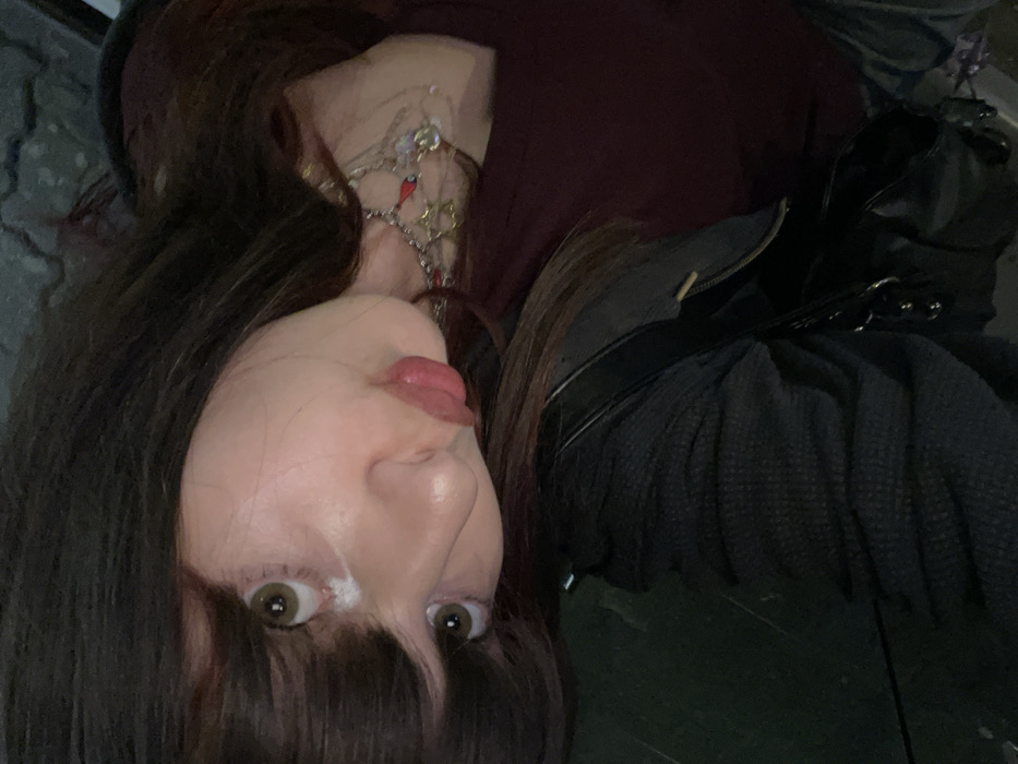 A woman is lying down facing the camera, sticking out her tongue. She has dark hair with bangs and brown eyes. She is wearing a maroon shirt and a silver necklace with colorful charms. A black leather jacket and dark sweater are draped over her.