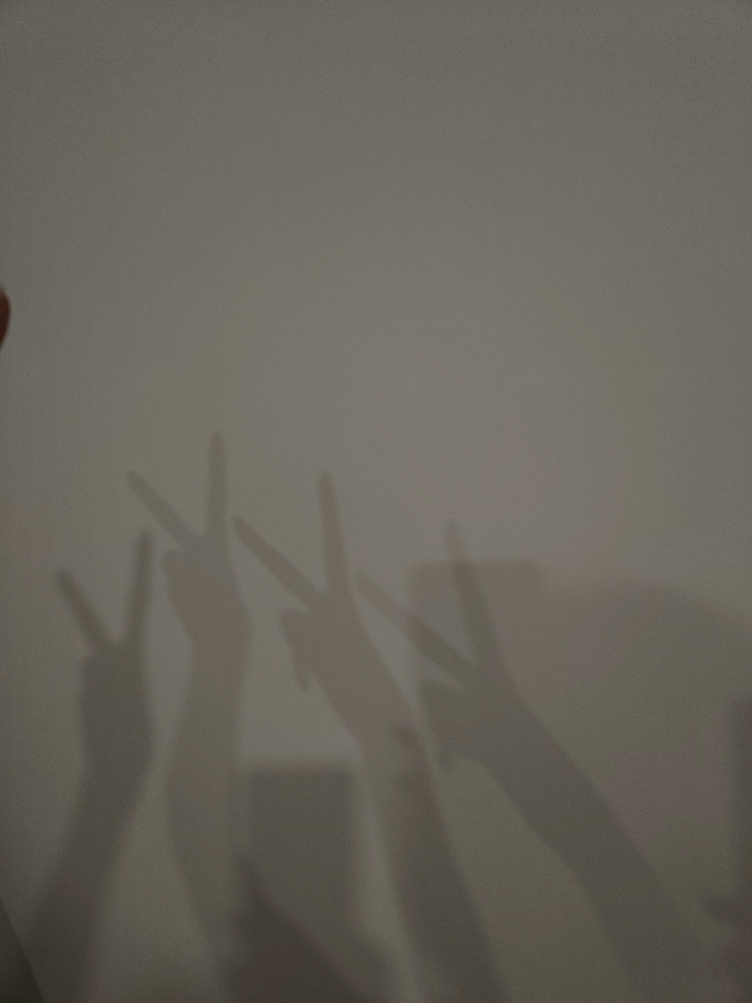 The image captures an indoor scene dominated by the shadows of four hands cast upon a plain, light-colored wall. Each hand is positioned to form a "peace" sign, with the index and middle fingers extended in a V-shape.
From left to right, the first hand shadow is partially visible along the frame's edge. The subsequent three hand shadows are fully displayed, showing clear outlines of the fingers and wrists. Notably, directly beneath the shadow of the second hand from the left, there is an additional distinct shadow of a small, rectangular object, which could be a mobile phone or a similar device.
The shadows are dark grey against the lighter background, indicating a single, unseen light source positioned to project these forms. The wall itself appears to be a neutral, untextured surface, possibly painted an off-white or light grey color.
The activity depicted is a playful interaction, likely involving multiple individuals posing their hands to create these specific shadows. The peace signs suggest a theme of camaraderie, victory, or a lighthearted moment. There are no specific location cues beyond an generic indoor setting, and no visible text is present in the image. The provided crops further highlight the wall's subtle texture and the clarity of the cast shadows, including the rectangular object's outline.