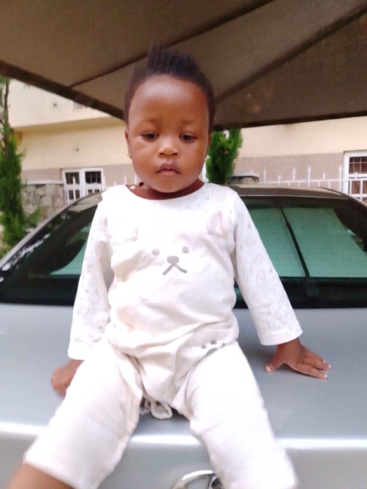 A young child sits on the hood of a silver car. The child is wearing a white outfit with a cartoon character on the chest. The child's legs are bent at the knees and their arms are resting on their lap. The car is parked outdoors, with a building and some trees in the background. The scene is well-lit, with natural light illuminating the child and the car. The overall tone of the image is casual and relaxed.