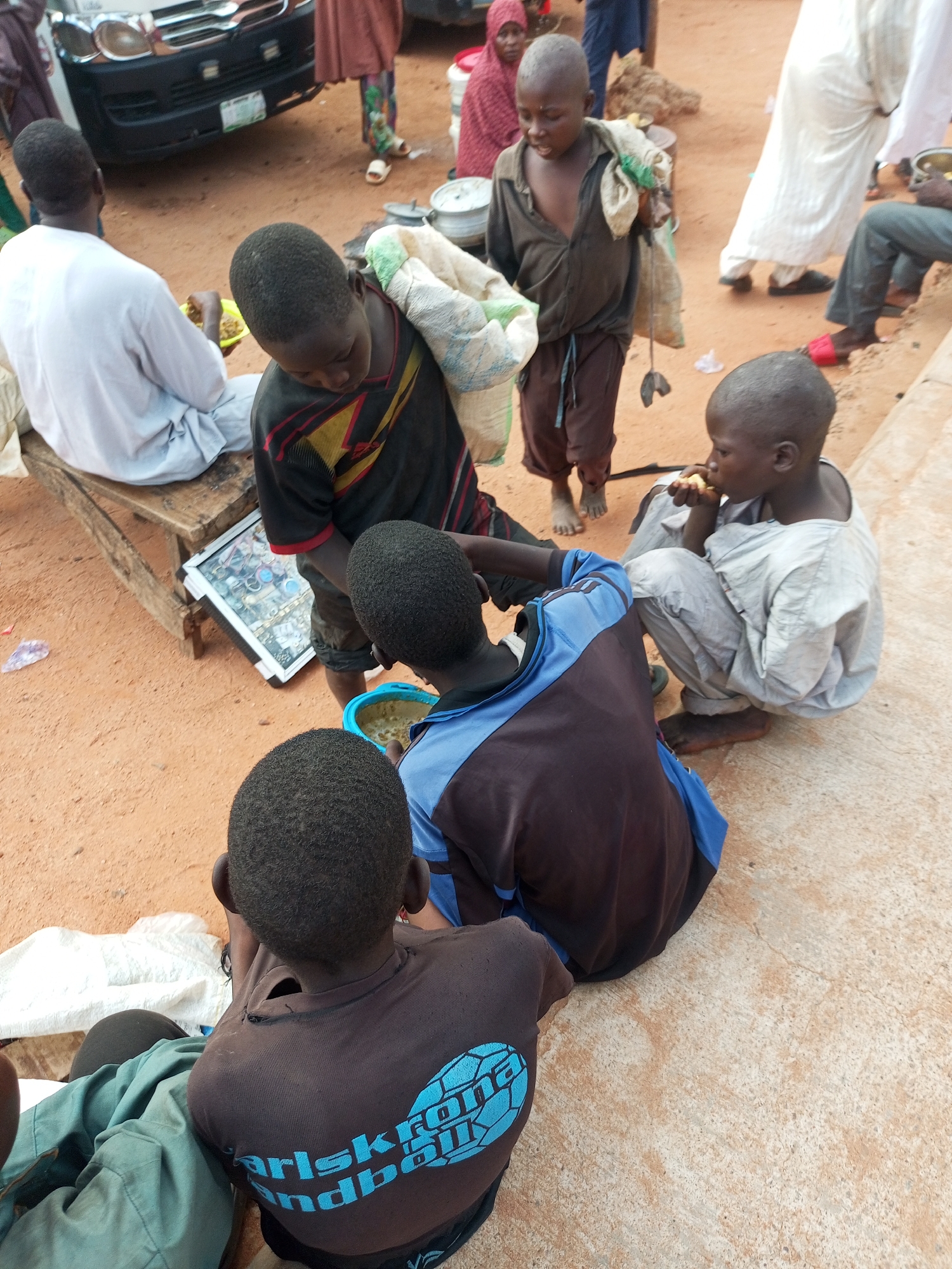 In the city of Gombe, Nigeria, a group of children are gathered on sandy ground near a road. In the foreground, two boys are sitting. One, wearing a brown shirt with blue lettering and a light blue collar, is facing away from the camera. The other boy, to his left, is partially obscured but appears to be wearing a green garment. To the right, a third boy in a light gray outfit is seated and eating something.
Behind them, a boy in a dark brown shirt and pants carries a large sack over his shoulder. He is standing near a wooden bench where another boy in a white robe is seated, holding a yellow bowl. A man or woman in a red headscarf and patterned pants stands further back.
In the background, a white vehicle is parked, and other individuals, including one in a white robe and red sandals, are visible. The environment is characterized by reddish-brown sand and worn concrete surfaces. The scene appears to be a casual gathering or market setting.