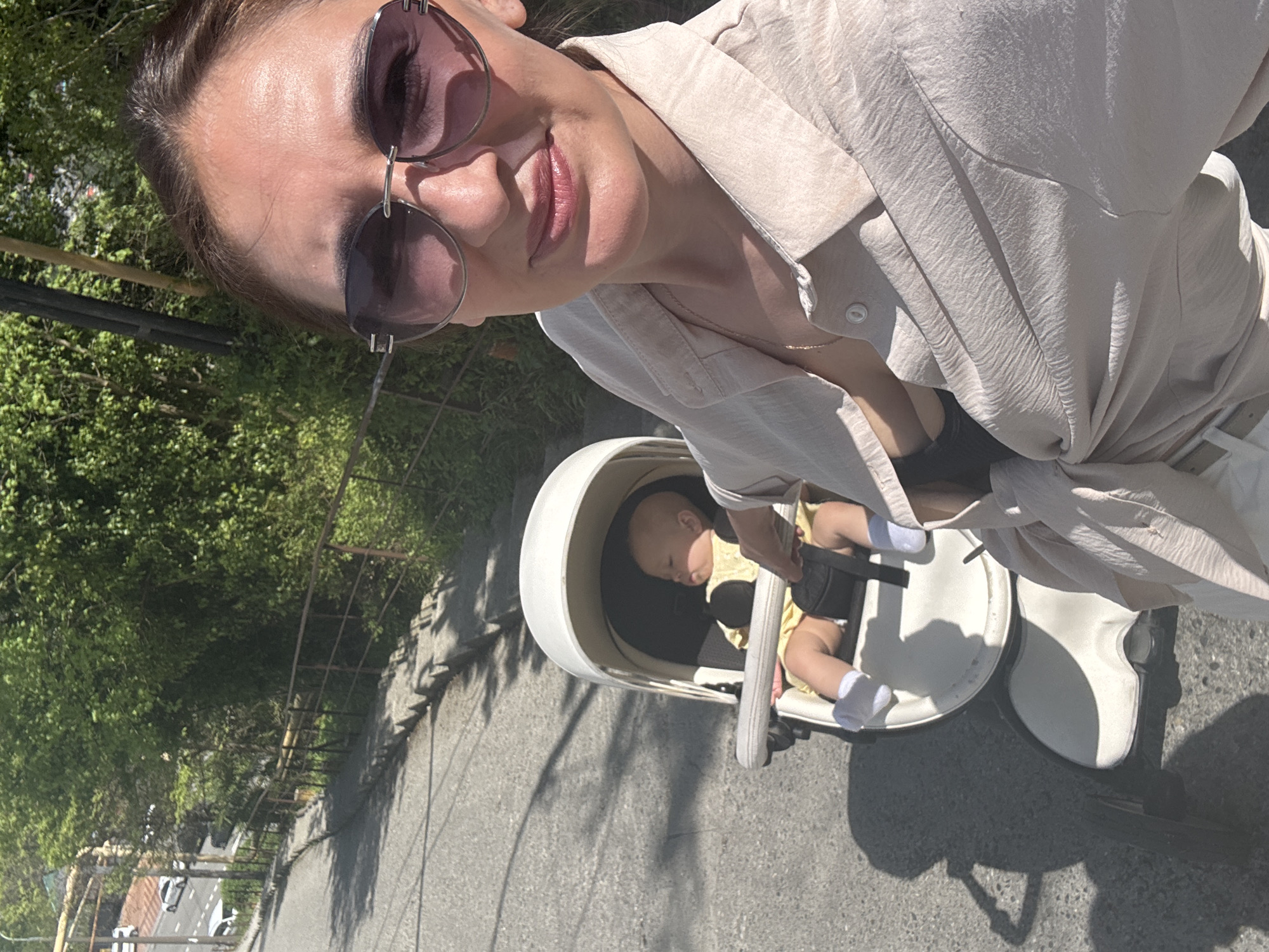 The image depicts a woman taking a selfie while standing next to a baby in a stroller. The woman is wearing sunglasses and a light-colored button-down shirt, partially unbuttoned. The baby is wearing white socks. They are on a street with a railing on one side and trees in the background. The location may be in Sochi, Russia, based on the urban environment.