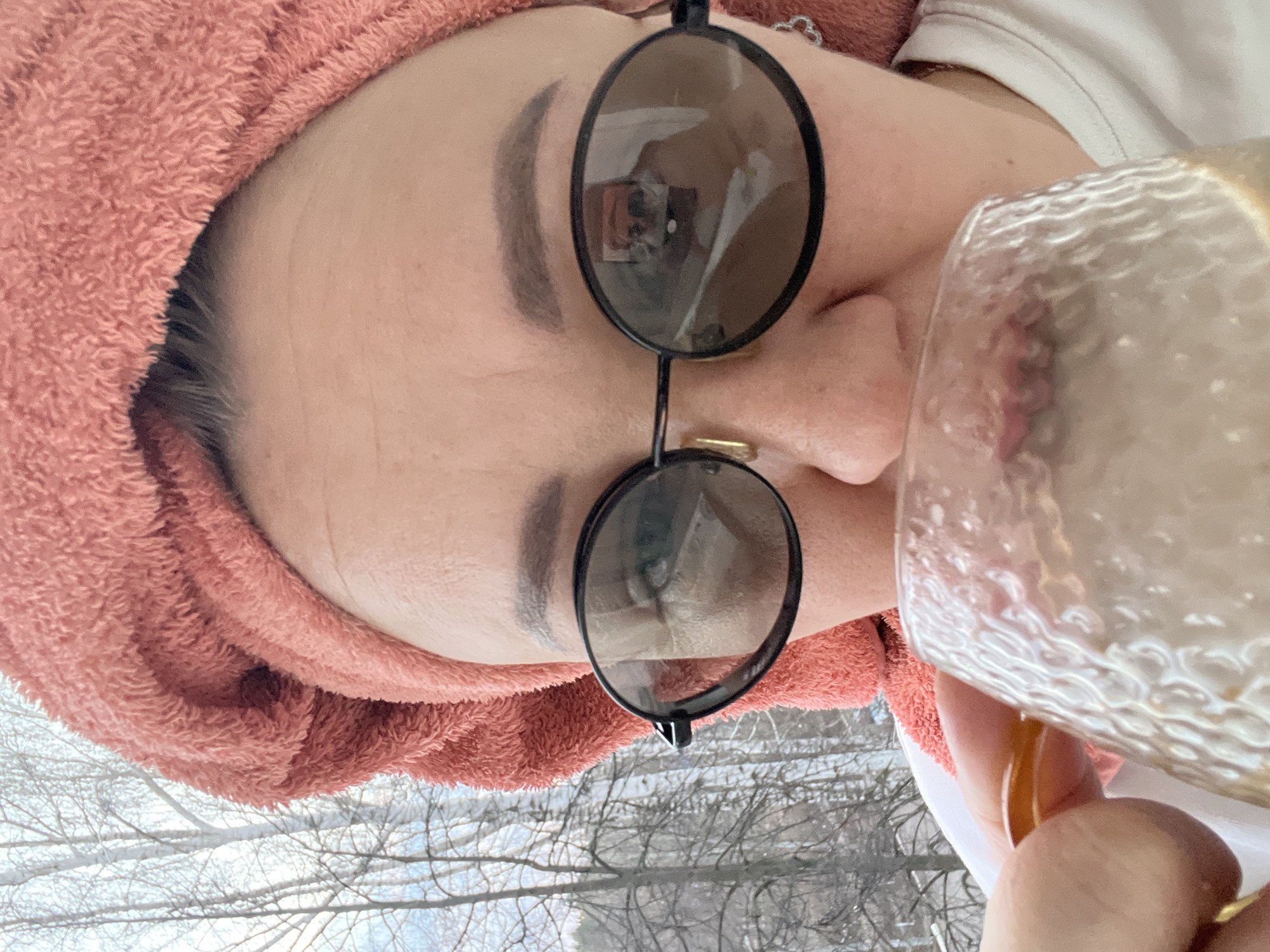 A person is wearing a peach-colored towel on their head and sunglasses with dark frames. The person is holding a clear glass with a golden liquid inside. Outside, there are trees without leaves, possibly in winter. The location is potentially Chaikovsky, Russia.