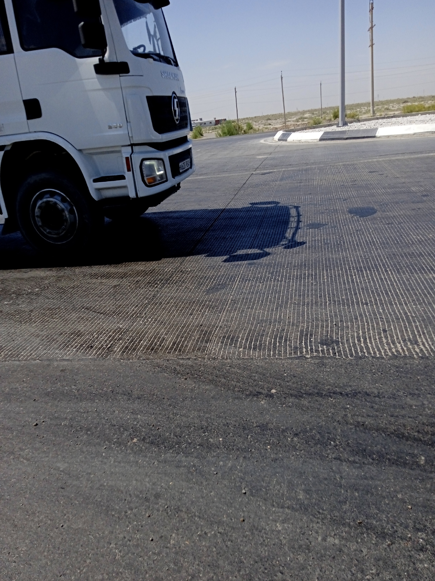 A white truck is parked on a paved road. The truck is facing right and its front wheel is visible. The road surface has a textured pattern of parallel lines. In the background, there is a sidewalk with a curb and some vegetation. The sky is clear and blue. The scene appears to be in an outdoor urban environment.