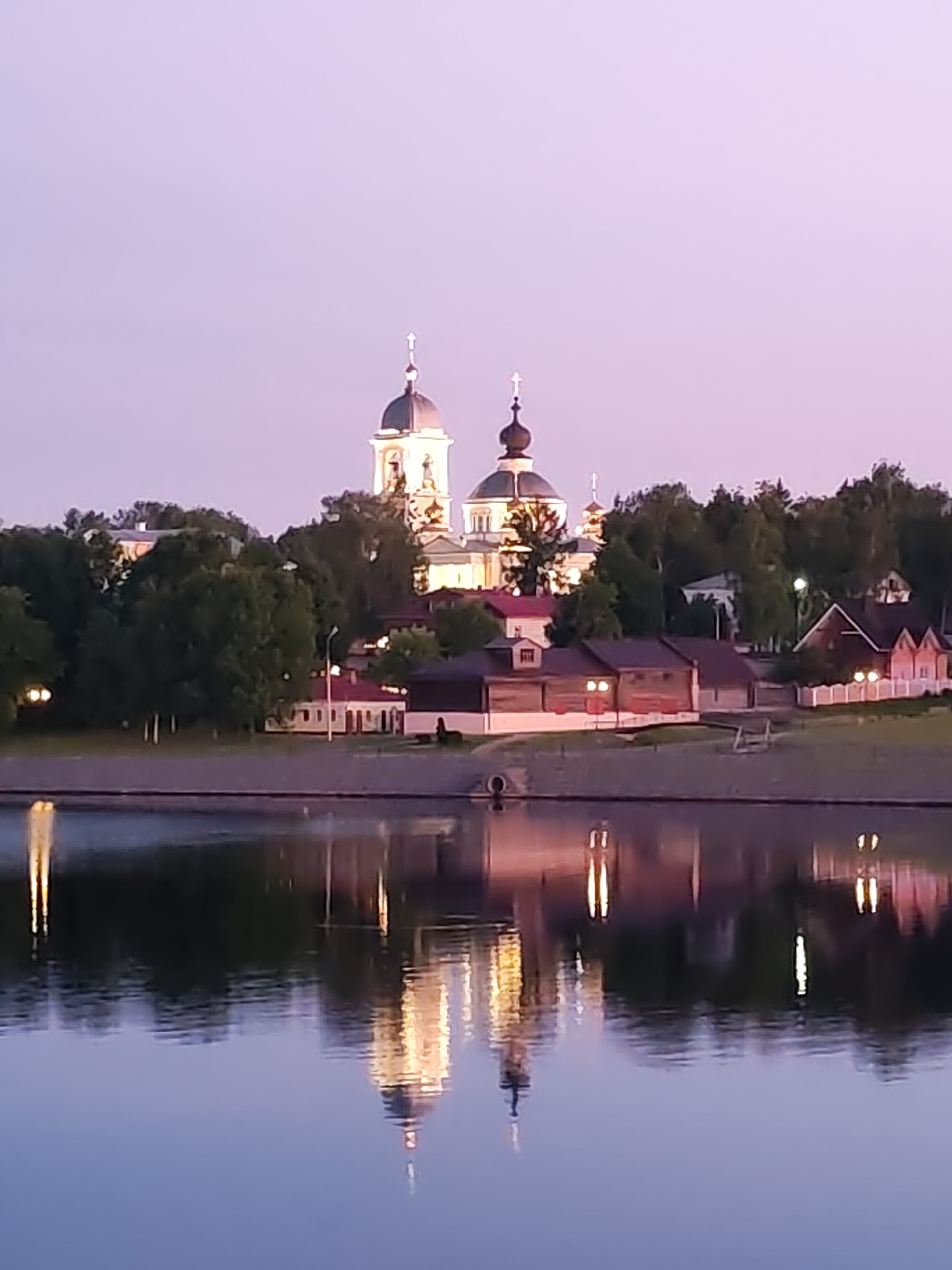 This detailed image captures a serene twilight scene of the city of Myshkin, Russia, along the bank of the Volga River. The upper portion of the frame features a clear sky displaying a gradient of light lavender to pale violet hues, indicative of dusk. Dominating the cityscape are two distinct Russian Orthodox churches, positioned centrally on a raised ground line. The left church exhibits lighter-colored domes, potentially white or silver, while the right church features darker, possibly black or deep green, onion-shaped domes. Both are prominently illuminated by artificial lighting, highlighting their architectural forms and the crosses atop their cupolas.
Below and around the churches, a cluster of smaller, two-story buildings with red or dark-colored roofs are nestled amongst a dense line of dark green deciduous trees. Several of these peripheral buildings display warm, yellow internal or external lighting. A notable building on the far right has a distinct pink exterior.
The lower half of the image is dominated by the calm, wide surface of the river, which acts as a reflective mirror for the illuminated structures and lights on the bank. The reflections are clear but slightly elongated and diffused by minor surface ripples. A dark, even strip of land or an embankment separates the water from the buildings, with a cylindrical drainage pipe visible at the waterline near the center. Artificial streetlights and building lights cast bright, distinct reflections across the water. The scene is devoid of any visible people, vehicles, or animals, presenting a tranquil and static view of the illuminated riverfront.