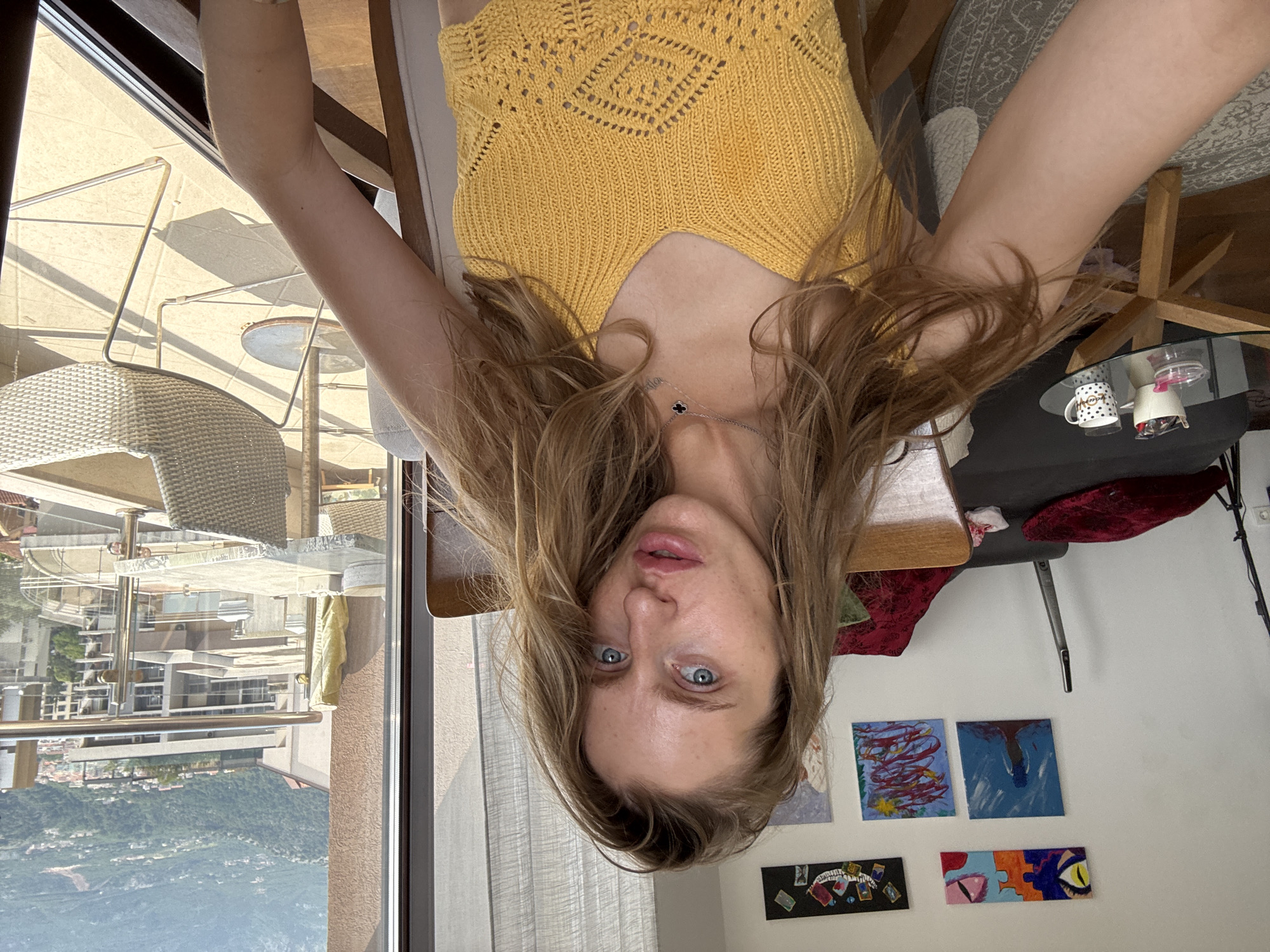 A young Caucasian woman with long, blonde, wavy hair is seated in a chair indoors, facing the camera. She is wearing a yellow knitted crop top with a V-neck and intricate patterns. Her eyes are open and directed at the lens, and her lips are slightly parted. A delicate necklace with a clover pendant is visible around her neck.
The background reveals an indoor setting with a wall adorned with several abstract paintings. To the left, a sofa with red cushions is partially visible, and a tall, slender cleaning device stands near it. A glass-top coffee table with a patterned mug and a small dish is also present.
Beyond the woman, a large window offers a view of a cityscape. The scene outside suggests a sunny day with buildings and a hilly landscape in the distance, indicative of a location like Budva, Montenegro, known for its coastal and mountainous terrain. An outdoor seating area with chairs and tables is also visible through the window. The overall atmosphere appears to be casual and relaxed.
