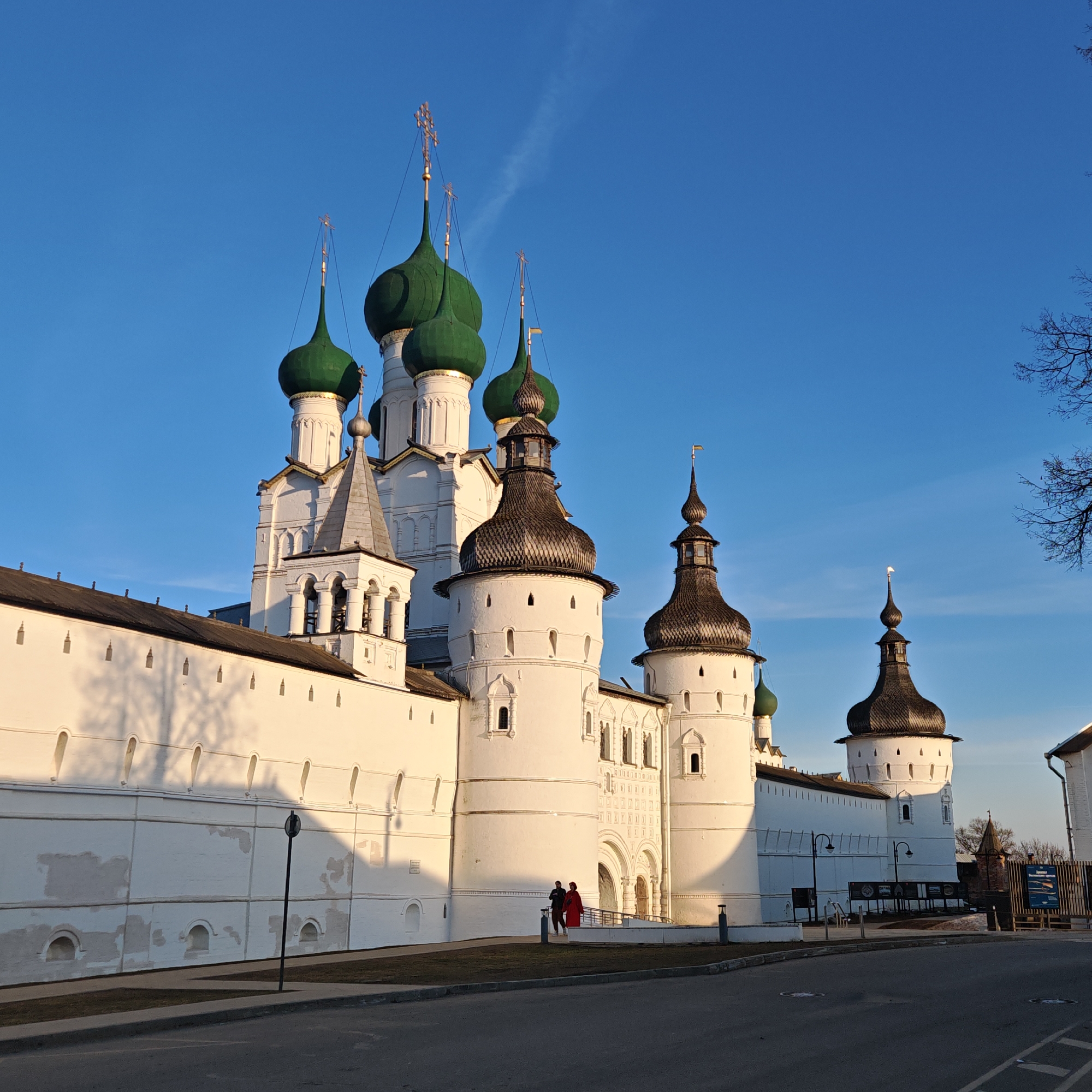 The image shows the Rostov Kremlin, located in Rostov, Russia. The complex is composed of white stone buildings, including tall walls with turrets, cathedrals with green domes, and a bell tower. The sky is clear and blue. In the foreground, there is a paved road and a small patch of grass. Two people are standing near the entrance to the Kremlin. The setting appears to be a sunny day.