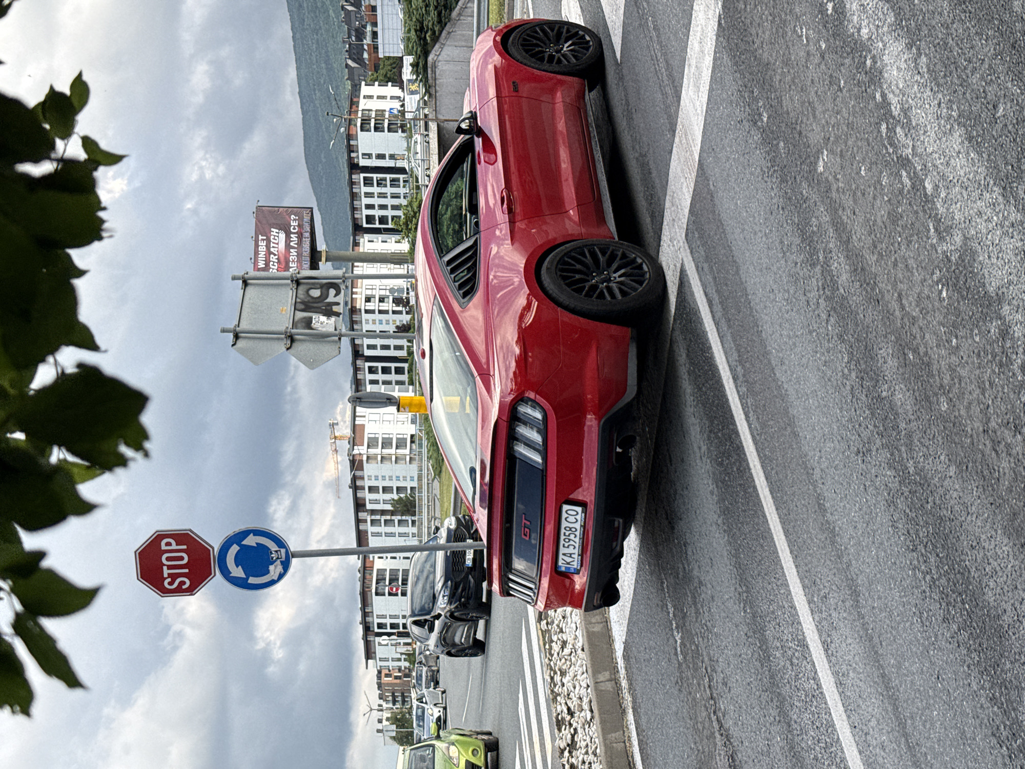 An outdoor, eye-level shot captures a vibrant red Ford Mustang GT on an urban street in Sofia, Bulgaria, under an overcast sky. The car is prominently featured, positioned on the right side of the road, appearing to be stopped or moving slowly near a crosswalk marked by white stripes. It is fitted with custom black multi-spoke wheels.
To the left of the Mustang, a black SUV or crossover vehicle is visible, followed by a line of other cars, including a partially visible lime green car. The background showcases several modern residential or commercial buildings, with distant mountains forming the horizon. A construction crane is also visible above the buildings. Lush green foliage from a tree frames the left side of the image.
Road signs are present on the left, including a red octagonal "STOP" sign and a blue circular sign indicating a roundabout or mandatory direction. A large billboard above the street displays "WINBET SCRATCH" in English, and the Bulgarian phrases "ЕЛЕЗИ ЛИ СЕ?" (Are you getting hooked?) and "РИСХ ОТ РАЗВИВАНЕ НА ЗАВИСИМОСТ" (Risk of developing addiction), a standard gambling disclaimer.
The rear of the red Mustang clearly displays "GT" branding and its Bulgarian license plate, "KA 5958 CO". A small "NISSAN" text is visible below the license plate, likely on a dealer frame. The road surface appears damp, suggesting recent rainfall or high humidity. The scene depicts a typical daytime street environment in a Bulgarian city.