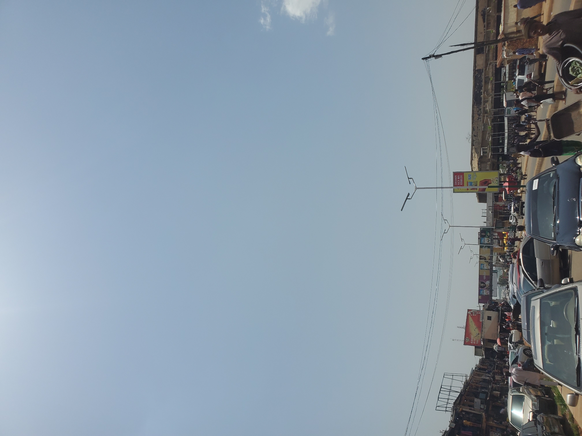 The image captures a bustling street scene in Jos, Nigeria, likely during daytime given the clear sky and bright sunlight. The perspective is from a street-level view, looking down a road lined with buildings and an array of parked and moving vehicles. A significant number of people are visible, suggesting a busy marketplace or thoroughfare. Some individuals are walking, while others appear to be engaged in commerce, with stalls or shops lining the sides of the road.
Objects prominently featured include various cars, some of which are parked bumper-to-bumper, indicating traffic congestion. There are also utility poles and overhead wires crisscrossing the sky, common in urban environments. Several billboards are visible, advertising products and services, one of which displays text in a foreign language, possibly related to a local brand or event. The buildings themselves appear to be a mix of commercial structures, some with multiple stories.
The overall impression is one of a lively and active urban environment. The weather appears to be clear and sunny, with a few wispy clouds in the sky. The scene conveys a sense of everyday life and economic activity in Jos, Nigeria, with the constant movement of people and vehicles. The orientation of the image is rotated, with the sky appearing on the left and the ground on the right.