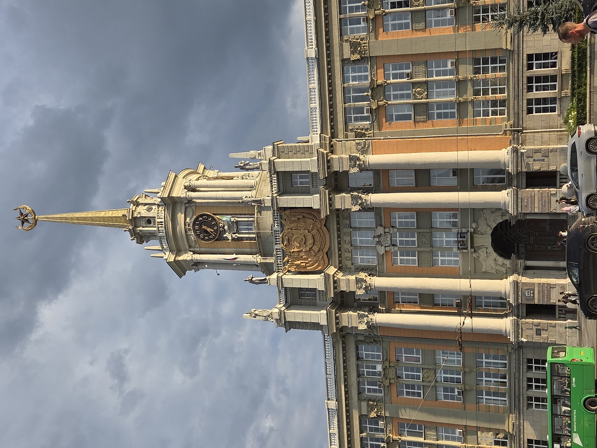 This image shows a view of the Yekaterinburg City Administration building, also known as "Gorsovet", located in Yekaterinburg, Russia. The imposing building features classical architectural elements, including columns, statues, and a large clock on its central tower, which is topped with a star. The facade is a mix of light and darker orange-toned stone. The sky above is partly cloudy with dramatic, dark clouds that suggest an approaching storm or a dramatic sunset.
In the foreground, a green bus with "ИНФРАСТРУКТУРА" and "ГТВК" written on its side is parked near the curb. Several cars are visible on the street, including a black sedan and a silver SUV. A few pedestrians are walking on the sidewalk in front of the building. A man is standing on the far right of the image, looking towards the right, and a small evergreen tree is visible to his left. The overall impression is one of grand Soviet-era architecture under a dynamic sky.