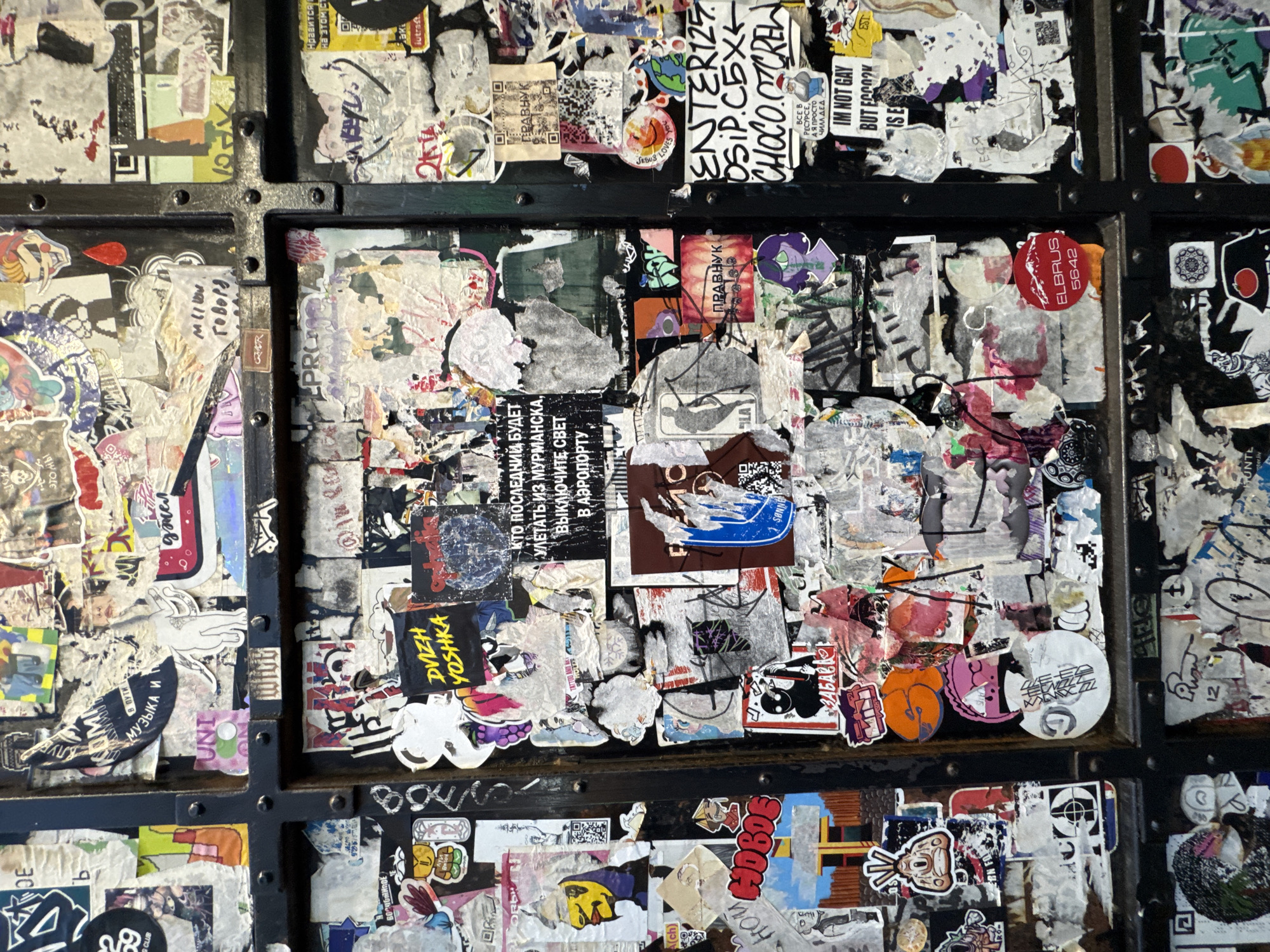 The image shows a densely packed collection of stickers and graffiti on a dark-colored surface, possibly a wall or structure in Moscow, Russia, based on the presence of Russian text. The scene appears to be a public space heavily marked with various forms of street art and messages. The objects include stickers, possibly posters, and other forms of adhesive art. The environment is urban, indicated by the graffiti and sticker accumulation, suggesting a high-traffic area in the city.