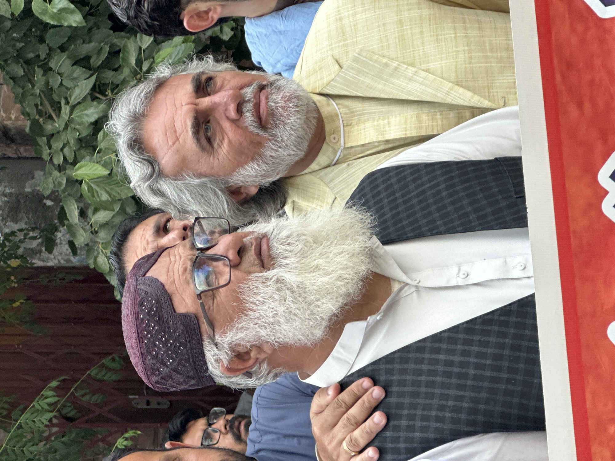 A group of men stand together in Quetta City Tehsil, Pakistan. The men have white beards. One man wears a burgundy prayer cap and glasses. He has a white shirt and a black and grey plaid vest. Another man wears a yellow jacket. The men stand next to a red sign with white text. Behind them is a green hedge.