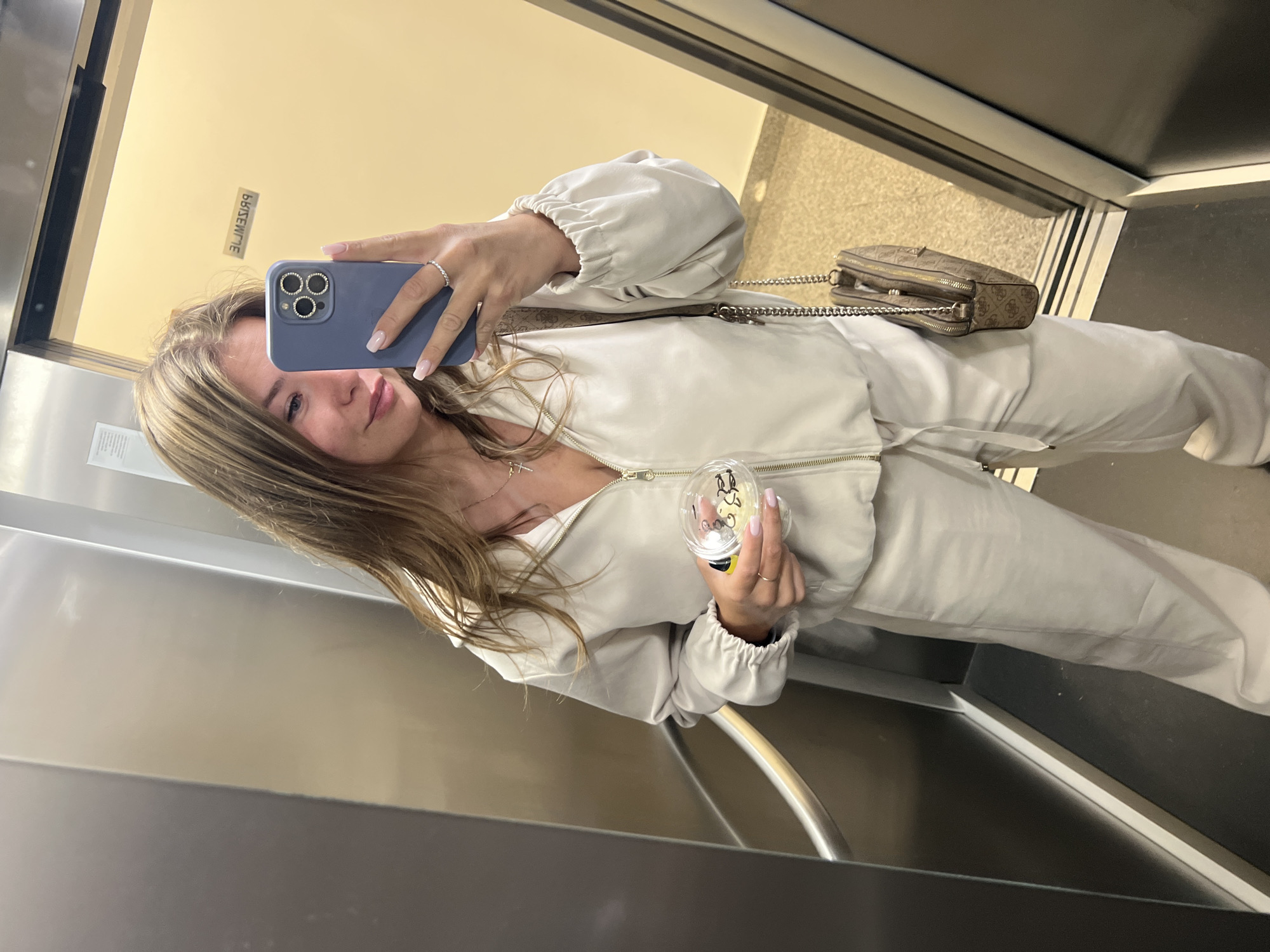 A young woman takes a selfie in what appears to be an elevator. She is holding a blue smartphone with a multi-lens camera in her left hand, and a clear plastic cup with yellow contents in her right hand. She wears a light-colored matching tracksuit, and a crossbody bag with a chain strap rests across her body. The interior of the elevator features stainless steel walls and a handrail. The elevator appears to be in a building in Budva, Montenegro, as suggested by the reversed sign visible on the wall. The floor outside the elevator is visible with neutral colored tile.