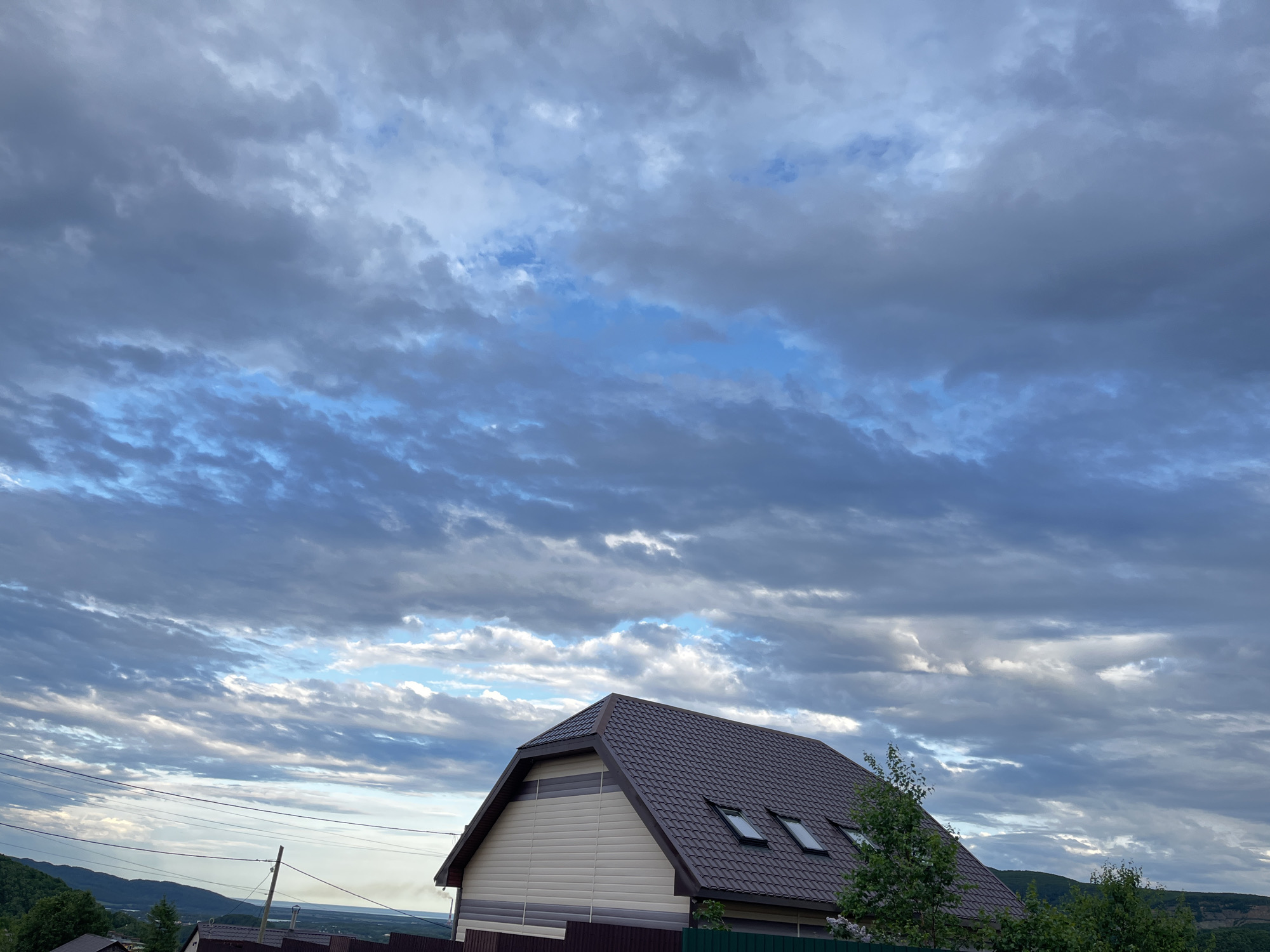 The image captures a suburban landscape under a dramatic sky, likely at dusk. A gable-roofed house with a dark brown shingled roof and beige siding dominates the foreground. Three dormer windows are visible on the roof. A dark brown fence runs along the front of the property. To the left, utility lines stretch across the frame, with a wooden pole supporting them. In the background, rolling green hills recede into the distance, culminating in a hazy horizon. The sky is filled with a complex pattern of grey and white clouds, interspersed with patches of bright blue sky. The lighting suggests the sun is low, casting a soft, diffused light on the scene. There are no people visible in the image. The location is identified as Petropavlovsk-Kamchatsky, Russia.