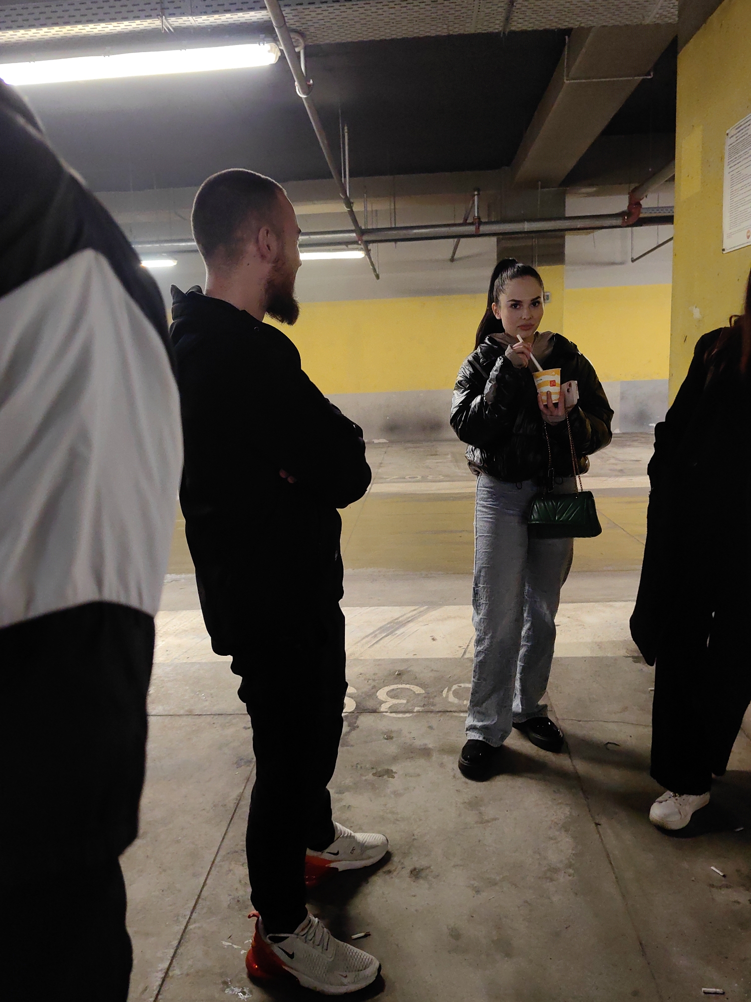 In an indoor parking garage in Sofia, Bulgaria, three individuals are observed. A man with a beard, wearing a black hoodie and white sneakers with red accents, stands with arms crossed. A woman in a black puffy jacket and light-wash jeans holds a beverage in a McDonald's cup and a green purse hangs from her shoulder. Part of a third person wearing a black jacket is visible. Cigarette butts are scattered on the concrete floor. The background shows a yellow wall and overhead pipes and lighting.