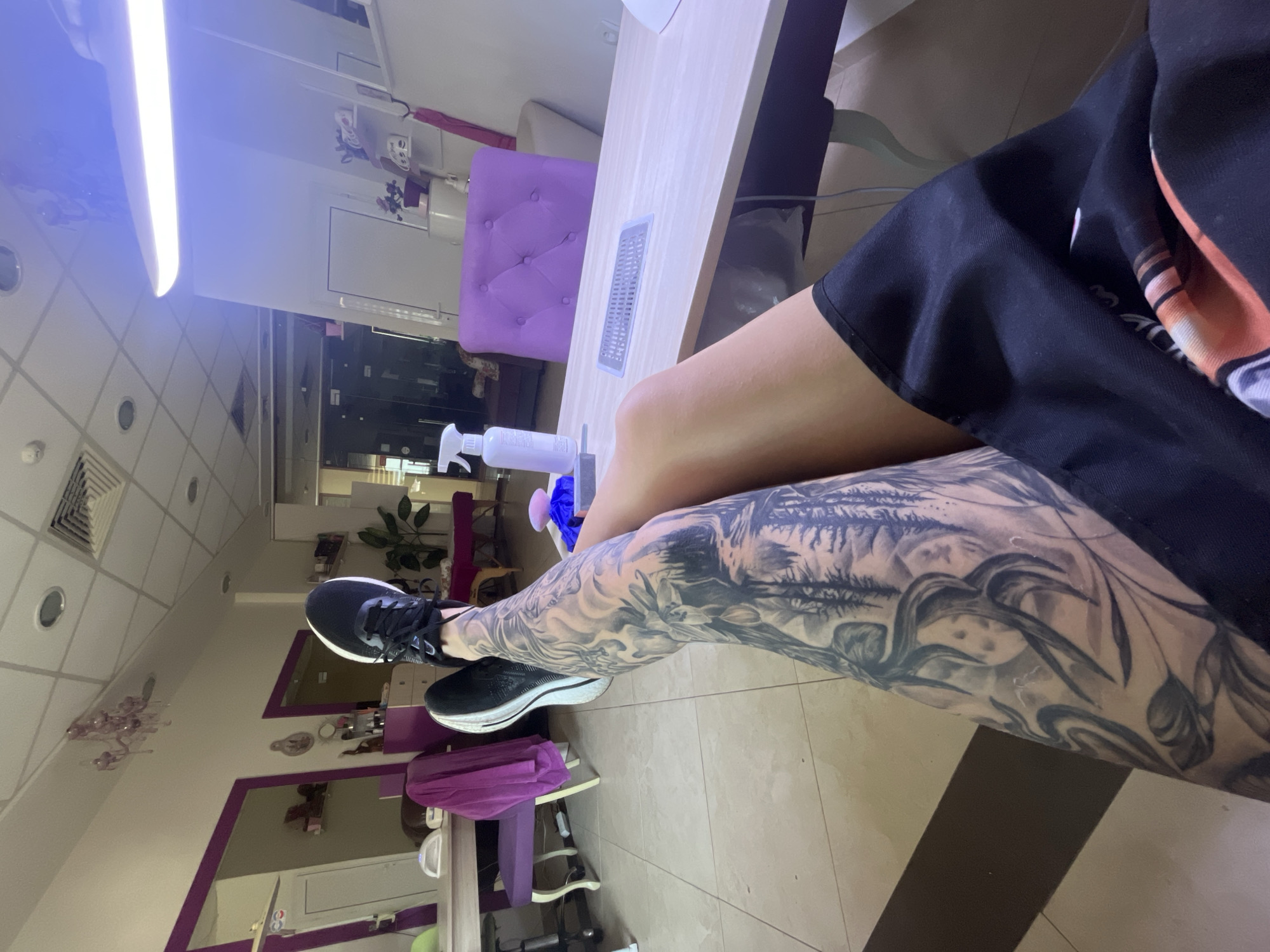 The image depicts a person sitting in what appears to be a beauty salon, possibly in Veliko Tarnovo, Bulgaria, given the context. The person is wearing black shorts and black sneakers with white soles. The person's left leg, which is prominently displayed, has a detailed black and gray tattoo. The person is seated at a manicure table. The manicure table has various items on it, including a spray bottle. A purple chair is nearby. The room has a tiled floor. There is a mirror with a purple frame.