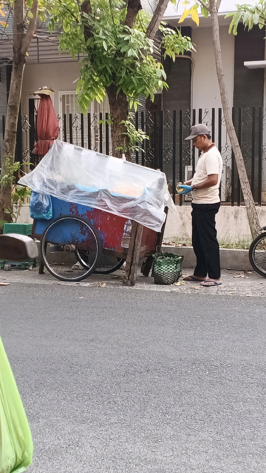 A man wearing a beige t-shirt, dark pants, a gray baseball cap, and blue latex gloves stands next to a food cart in Semarang, Indonesia. The cart is covered with a clear plastic sheet and has a red and blue painted body with two wheels. A green woven basket sits on the ground next to the cart, containing food scraps. The man appears to be working, possibly preparing or serving food. The scene is set on a street with trees and buildings in the background.