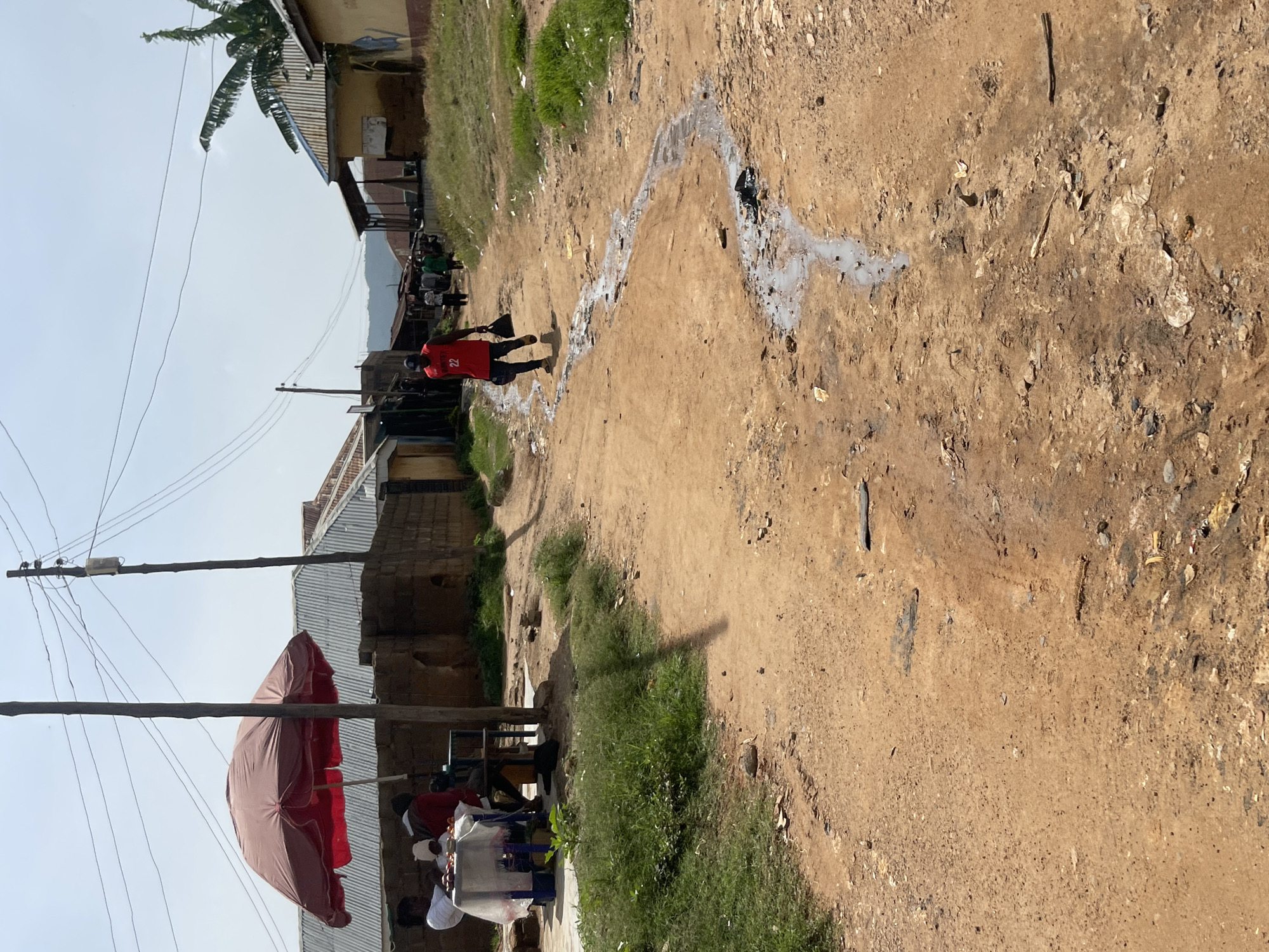 The image depicts a street scene in Jos, Nigeria. A dirt road, marked by a flowing channel of water and debris, runs through the center of the frame. On the left side of the road, a market stall is set up under a large red umbrella, with several people gathered around a table. Further down the road, a person wearing a red shirt with the number "22" on it is walking away from the camera, carrying a black bag. Several other individuals are visible walking in the distance, and buildings with corrugated metal roofs line both sides of the street. The environment appears to be an urban or semi-urban area, with evidence of both residential structures and commercial activity.