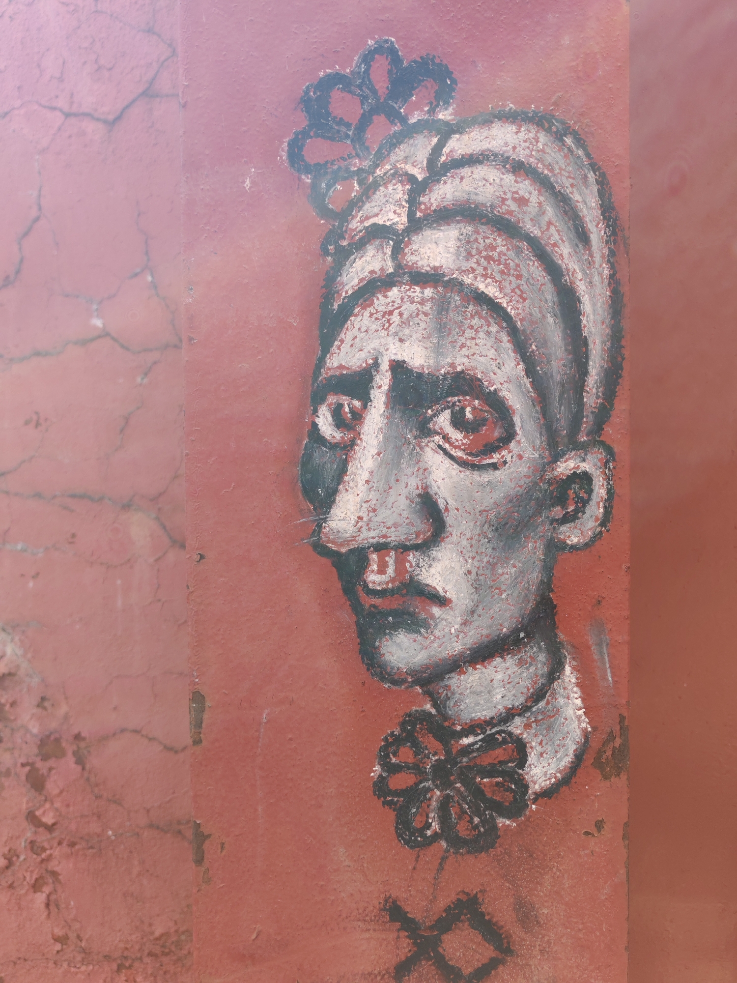 The image is a medium shot of a mural painted on a cracked, reddish-pink wall. The mural depicts a stylized portrait of a person's head and shoulders. The face is painted in shades of gray, white, and black, with red accents around the eyes and lips. The subject has a prominent nose, a thin mustache, and is wearing a turban-like headdress adorned with a black, flower-like embellishment. A black, bow-tie shape is visible at the neck. Below the neck, a black symbol resembling a stylized "X" or two crossed lines is painted. The wall shows signs of wear and tear, with visible cracks and peeling paint, particularly on the left side of the image. The overall aesthetic is somewhat primitive and expressive. There is no indication of the specific location in Moscow, Russia.