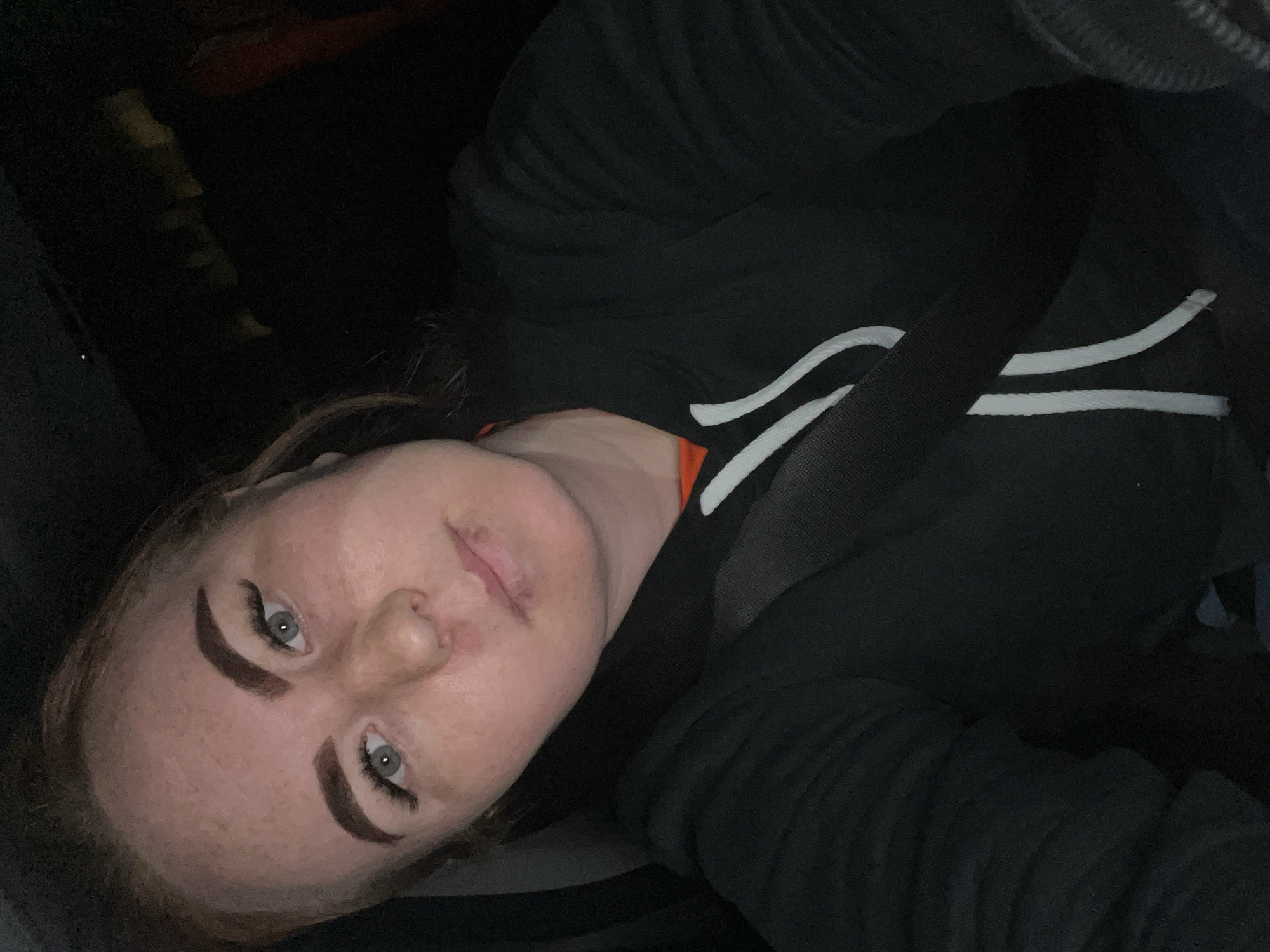 A woman with fair skin and red hair is seated in what appears to be a car, with a seatbelt fastened across her chest. She is wearing a black hooded jacket with white stripes on the sleeves. Her eyes are light-colored, and her eyebrows are darkly filled in. The surrounding environment is dark, suggesting it might be nighttime or a dimly lit interior. There is no visible information to suggest the city of Noviy, Russia is her location.