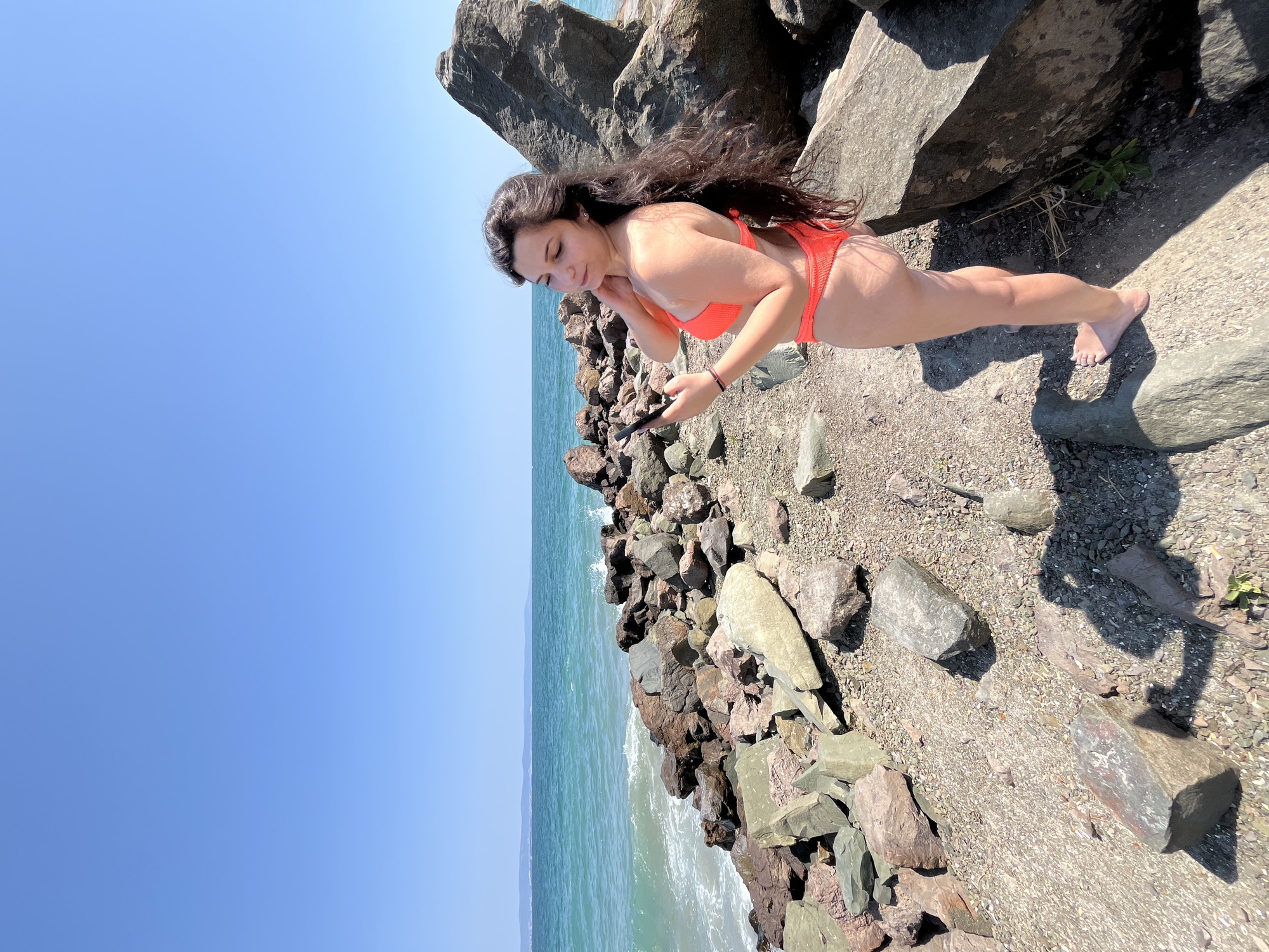 A young woman in an orange bikini stands on a rocky shore near the sea. She is looking at her phone and her long, dark hair is blowing in the wind. The sea is turquoise and calm, with waves gently lapping at the shore. The sky is clear and blue. The rocks are varied in size and color, with some covered in moss and others bare. The location is the rocky shore of Pomorie, Bulgaria, specifically the area near the salt pans which are known for their mineral-rich mud.