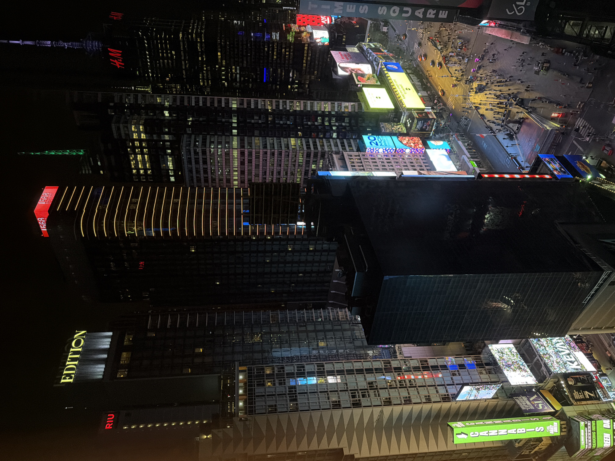The image captures an aerial view of Times Square in New York City at night. The scene is dominated by illuminated skyscrapers, including the Riu Hotel, EDITION Hotel, and a building with the "HM" logo. Bright digital billboards advertise a variety of products and services. People are visible in the lower-right corner of the image, moving through the streets.