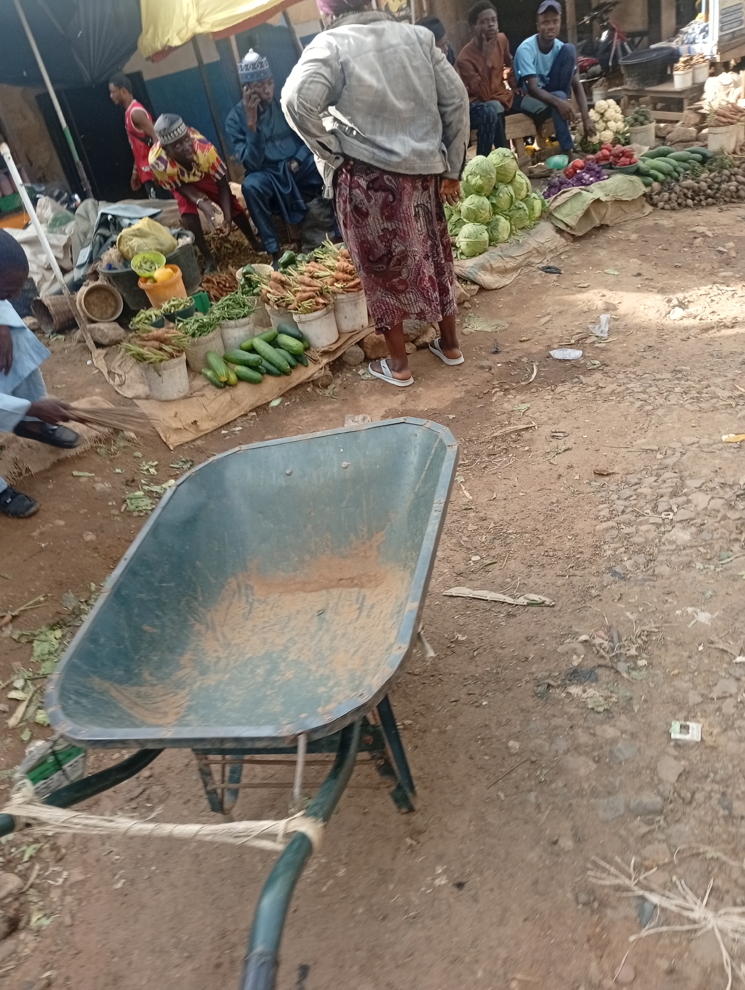 A busy outdoor market scene in Rafin Par, Nigeria. Several people are present, including men in traditional attire and a woman in a patterned skirt and jacket. The setting is a dirt ground with various stalls or displays of produce.
Objects prominently featured include a green wheelbarrow in the foreground, appearing dusty and well-used. Several buckets and baskets are filled with fresh vegetables, such as carrots and cucumbers, arranged for sale. Piles of cabbages and other produce are visible in the background. Some individuals are engaged in activities related to the market, such as a person sweeping with a broom and others standing or sitting near the produce displays.
The time of day appears to be daytime, with ample sunlight casting shadows, suggesting it could be mid-morning or afternoon. The weather appears to be warm, typical of a Nigerian market. The overall atmosphere suggests a lively and active marketplace. No visible text is present in the image. The scene depicts the everyday commerce and community interaction characteristic of a local market.