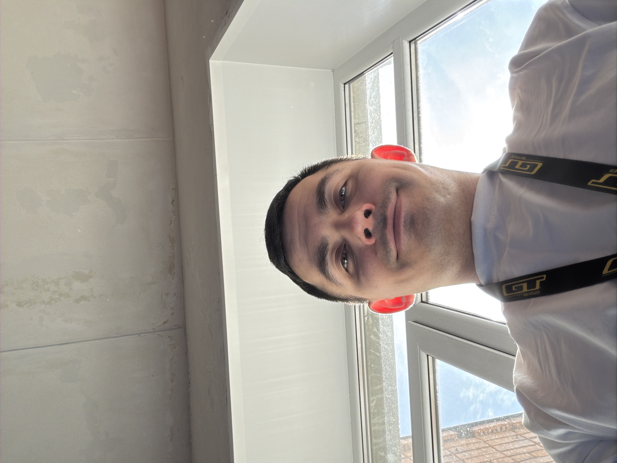 The photograph depicts a man indoors, positioned near a window. The environment appears to be a room with white walls. The window looks out onto a view of a building, perhaps indicating an urban or suburban setting. The man is wearing a white shirt and has black straps across his shoulder with the words "GT Bicycles" on them. No information allows the image to be linked to the city of Izhevsk, Russia.