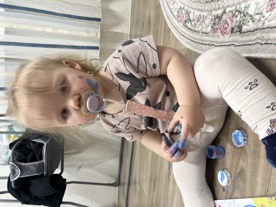 A young child with blonde hair, wearing a bunny-print shirt and white leggings with paw prints, sits on a wooden floor. The child has blue paint smeared on their face and hands, with several open paint lids nearby. They have a pacifier in their mouth with a patterned strap. A chair with a dark jacket hanging on it is visible on the left side, and a floral rug is partially seen in the background. A striped curtain hangs behind the chair.