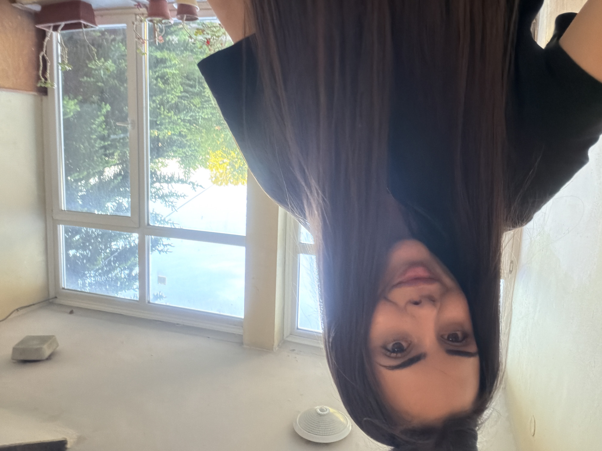 A woman with long, dark brown hair, wearing a black garment, appears upside down in the frame. Her face is prominently visible, with focus on her eyes, nose, and mouth. The background features a room with large windows showing trees and outdoor light. Plant pots are visible above the window. The floor is a light color and contains a few objects, including a square block and a small, circular white item. The location might be a balcony or a sunroom, judging by the windows and plants, however it's difficult to confirm if it's in Sofia, Bulgaria without more context.