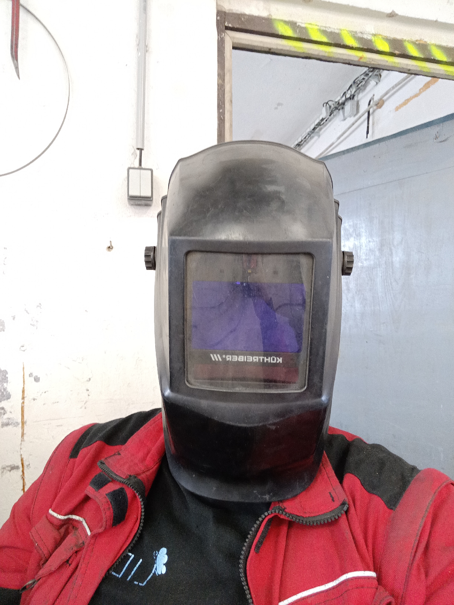A close-up, eye-level shot captures a person wearing a black auto-darkening welding helmet, which completely obscures their face. The helmet's tinted screen reflects its surroundings, revealing mirrored text that appears to be "KOHLBEIBER." and "ШІ.B.". The individual is dressed in a red and black work jacket, featuring visible zippers and contrasting panels, worn over a black t-shirt. On the t-shirt's chest, a light blue graphic or logo resembling stylized text (possibly "נורן") alongside a small abstract shape like a butterfly or moth is visible.
The setting appears to be an industrial workshop or utility space, located in Dalovy, Czechia. The background features a light-colored, possibly white, wall that shows signs of wear, scuffs, and minor peeling paint. A grey electrical conduit runs vertically on the wall, connecting to a small rectangular light switch or electrical box. A portion of a white circular object with a red and grey outline is partially visible on the left wall, possibly a clock or a sign. To the right and further back, a rectangular opening, likely a doorway or window, is framed by a raw concrete or wooden structure. The top edge of this opening is marked with distinctive yellow and black hazard stripes. Through this opening, more exposed wiring and conduits can be seen on the ceiling of an adjacent room. The presence of the welding helmet and work attire suggests the person is either preparing for or engaged in tasks such as welding or other manual labor within this industrial environment. The time of day is not discernible.