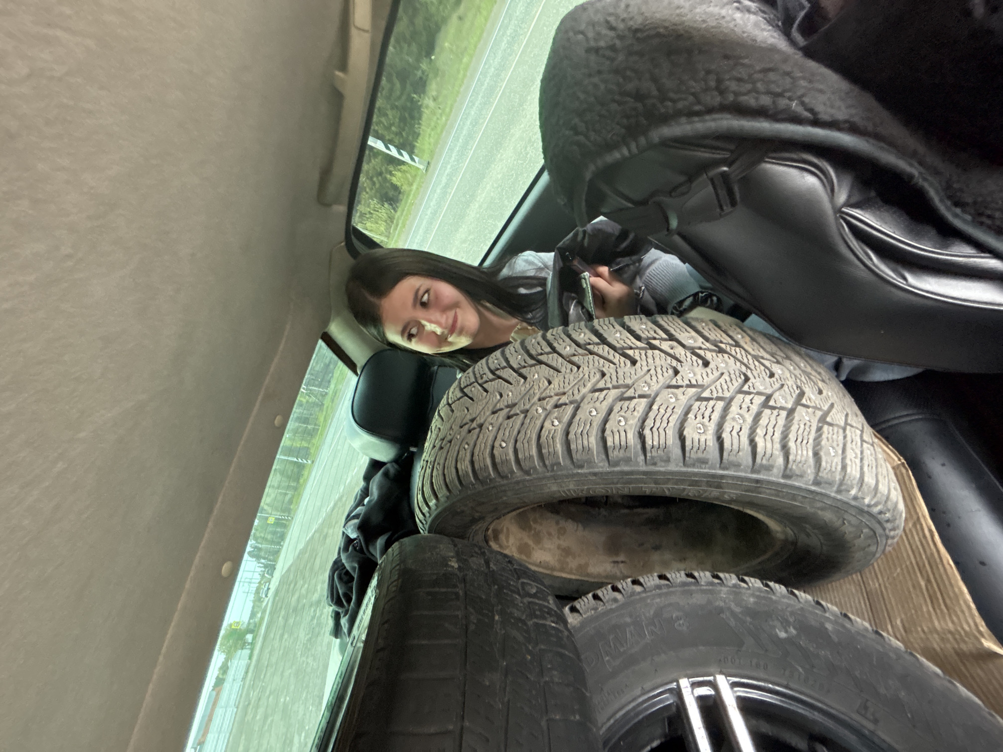 A young woman is in the back of a car in Chaikovsky, Russia. The trunk is filled with tires, and a leather coat is visible in the upper right corner of the frame. The tires are stacked on top of each other, with one tire having metal studs. The woman is looking towards the camera with a slight smile. Outside the car window, a street is visible.