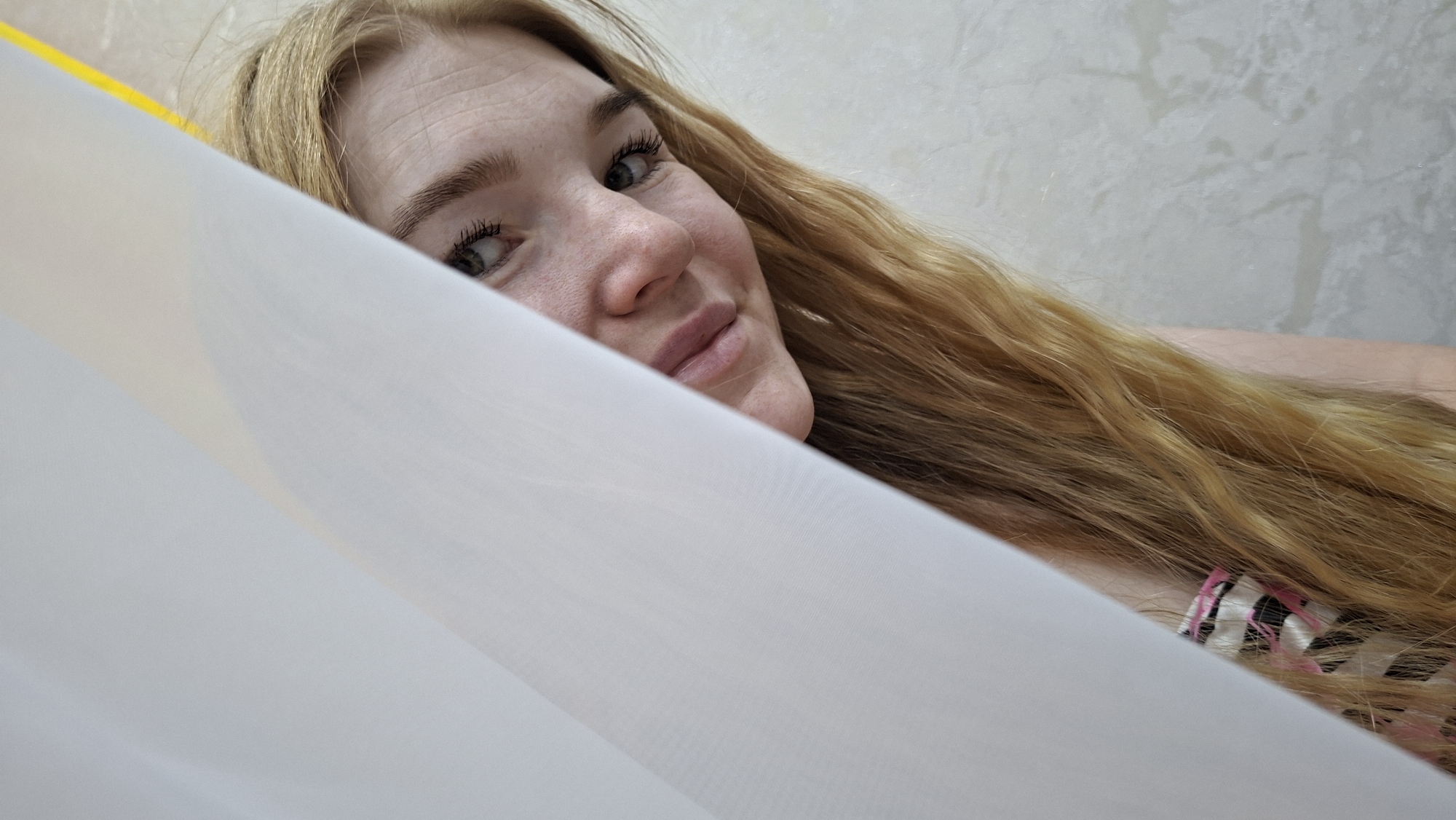 The image provides a close-up view of a young woman with long, flowing blonde hair, playfully peeking from behind a white, translucent fabric that occupies the foreground. Her head is tilted, and she looks directly at the viewer with a soft, engaging smile. Her light-colored eyes, possibly green or grey, are framed by dark eyelashes. The right side of her face, including her nose and full lips, is clearly visible, conveying a sense of warmth and approachability.
Her wavy, golden-blonde hair cascades over her shoulder, beneath which a glimpse of a patterned garment with pink and black stripes can be seen on her arm or upper body. The background consists of a light-colored wall with a subtle, textured pattern, suggesting an indoor setting. The lighting appears soft and natural, indicating a daytime scene. No specific time of day or weather conditions are discernible, other than an indoor environment. There is no visible text in the image. The overall mood is intimate and gentle, capturing a moment of playful interaction. This scene takes place indoors, within the context of Chaikovsky, Russia.