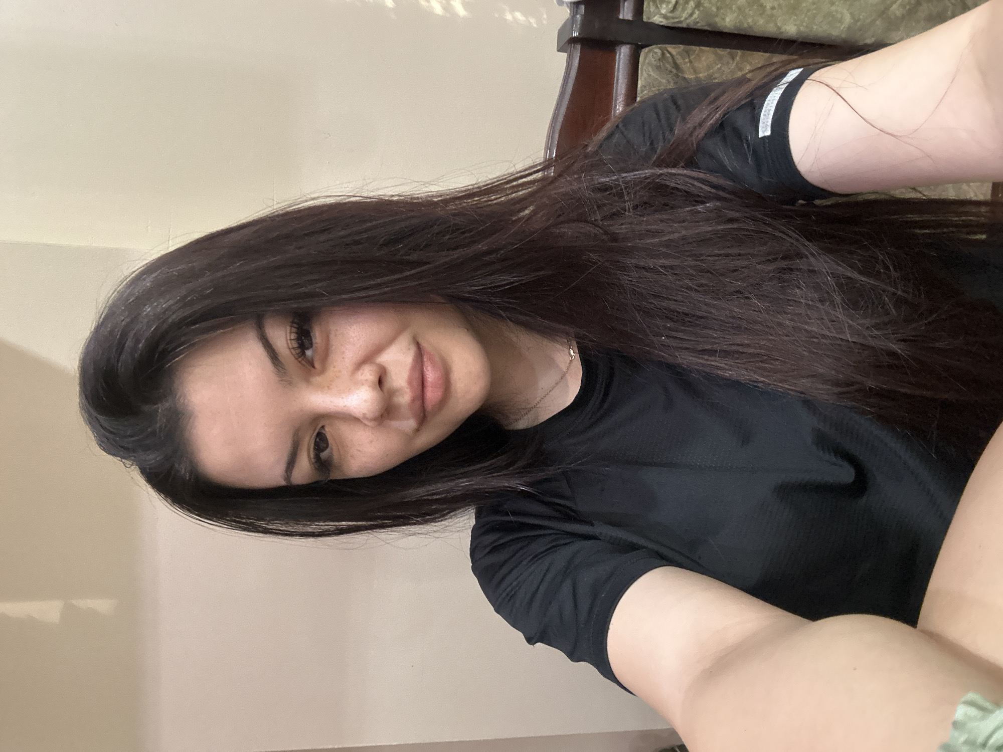 The image shows a young woman with long, dark brown hair and fair skin with freckles. She is wearing a black t-shirt and is looking towards the camera. The background appears to be an indoor setting with a beige-colored wall. A portion of a wooden chair with green upholstery is visible on the right side of the image. The lighting suggests it is daytime, possibly indoors with natural light from a window. The image quality is typical of a casual selfie. The specific location in the city of Sliven, Bulgaria, cannot be determined from the visual information provided.