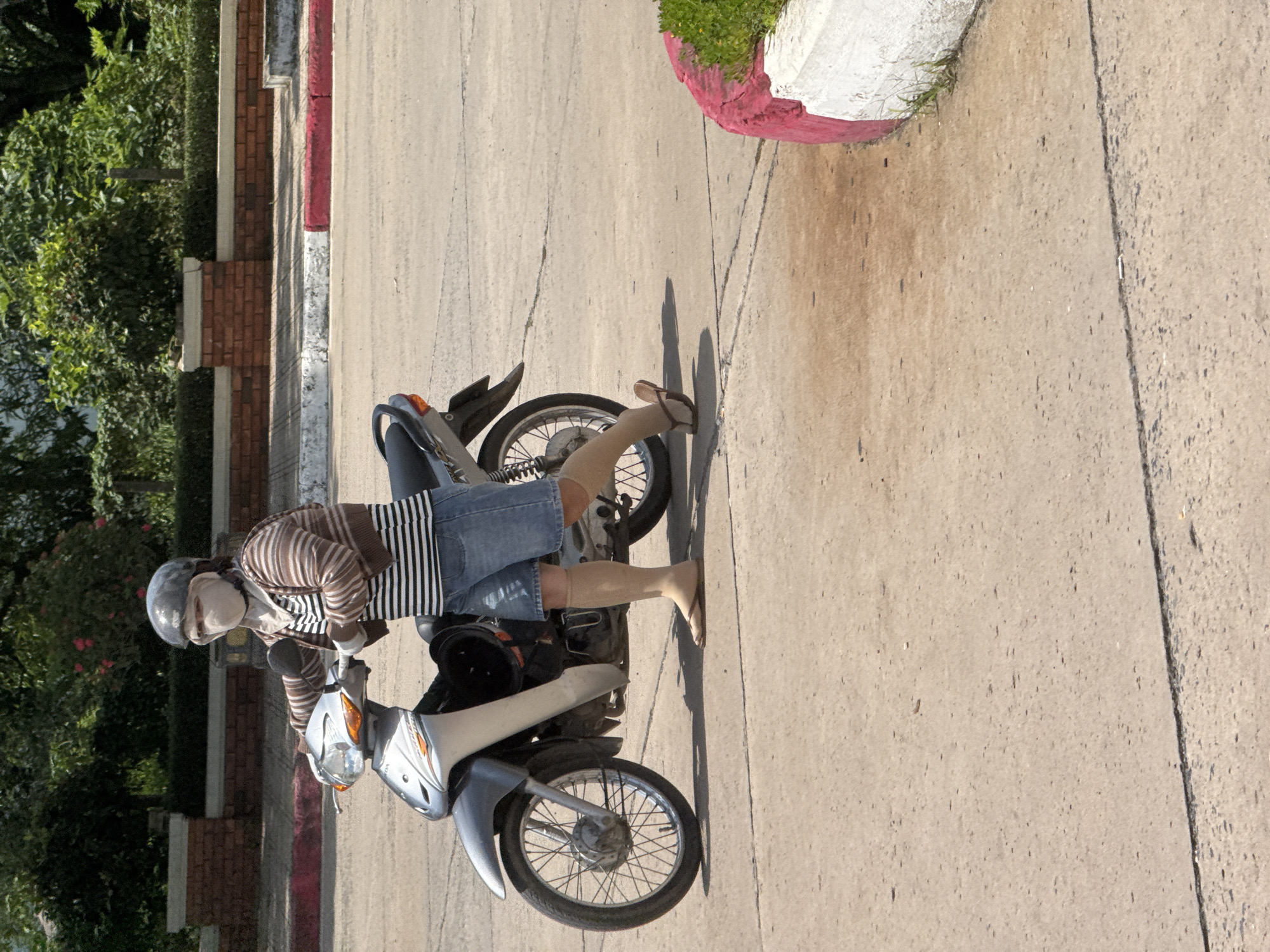 A person wearing a helmet, face mask, and a striped long-sleeved shirt is standing next to a silver motorcycle on a paved road. They are also wearing denim shorts and light-colored socks with flip-flops. The motorcycle's front wheel is turned to the left, and the person is holding onto the handlebars. In the background, there is a brick wall with a hedge and trees. The scene appears to be in an outdoor, urban environment, likely in Ben Cau, Vietnam, given the context.