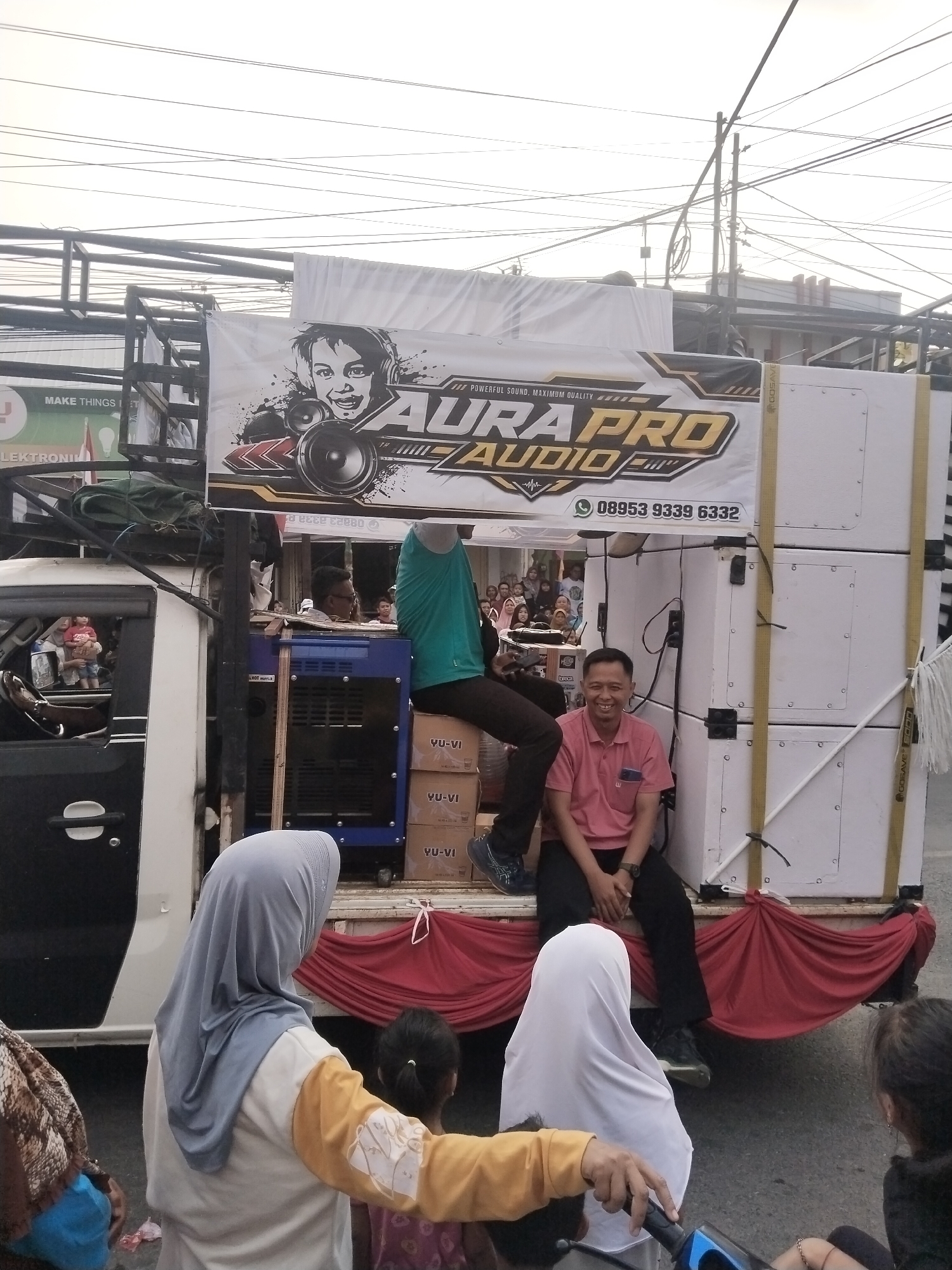 A low-angle, medium shot captures a flatbed truck parked on a street in Balapulang Kulon, Indonesia. The truck is laden with large, white speaker cabinets stacked vertically and a blue generator. A banner with the text "AURA PRO AUDIO" and a cartoon illustration of a child wearing headphones is prominently displayed on the side of the truck. Two men are visible on the truck; one, wearing a pink polo shirt and black pants, is seated and smiling, while another, wearing a teal tank top and black pants, is partially obscured. Several people, including women wearing hijabs, are gathered around the truck, some looking towards it. The background shows buildings and a tangled web of overhead electrical wires, typical of an urban Indonesian streetscape. The overall scene suggests a mobile sound system setup, possibly for an event or market.