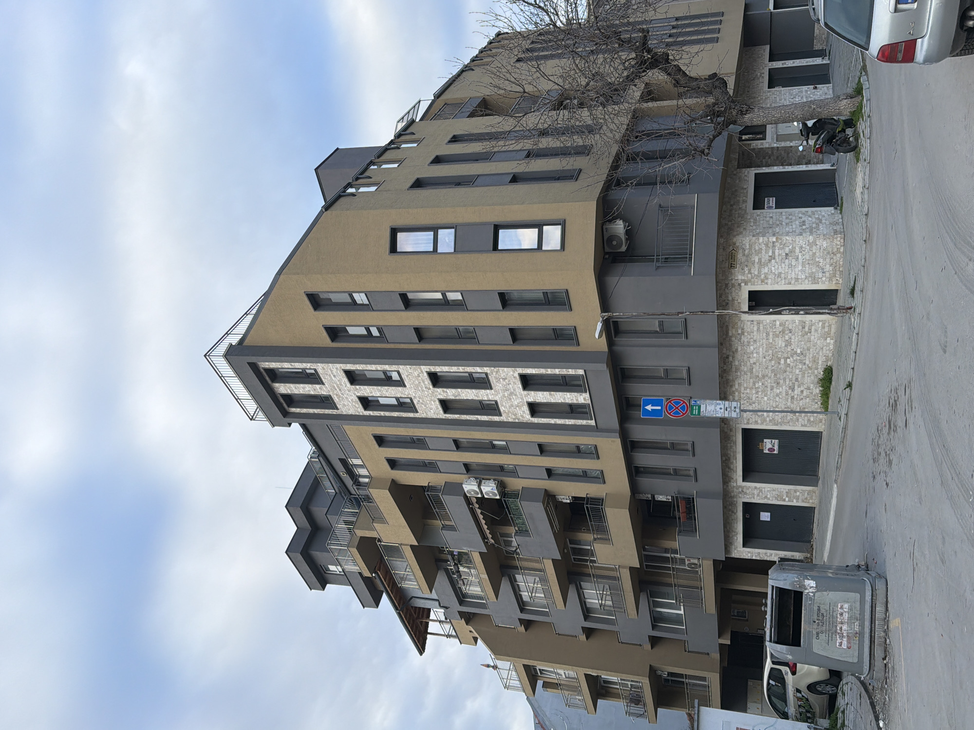 The image shows a modern apartment building in Varna, Bulgaria. The building has a unique architectural design, with alternating beige and gray sections and recessed balconies. A bare tree is visible in front of the building. A street sign indicating traffic regulations is present on the sidewalk, near the building. A parked motorbike, a gray metal trash bin, and a white car are visible along the street. The sky is overcast.