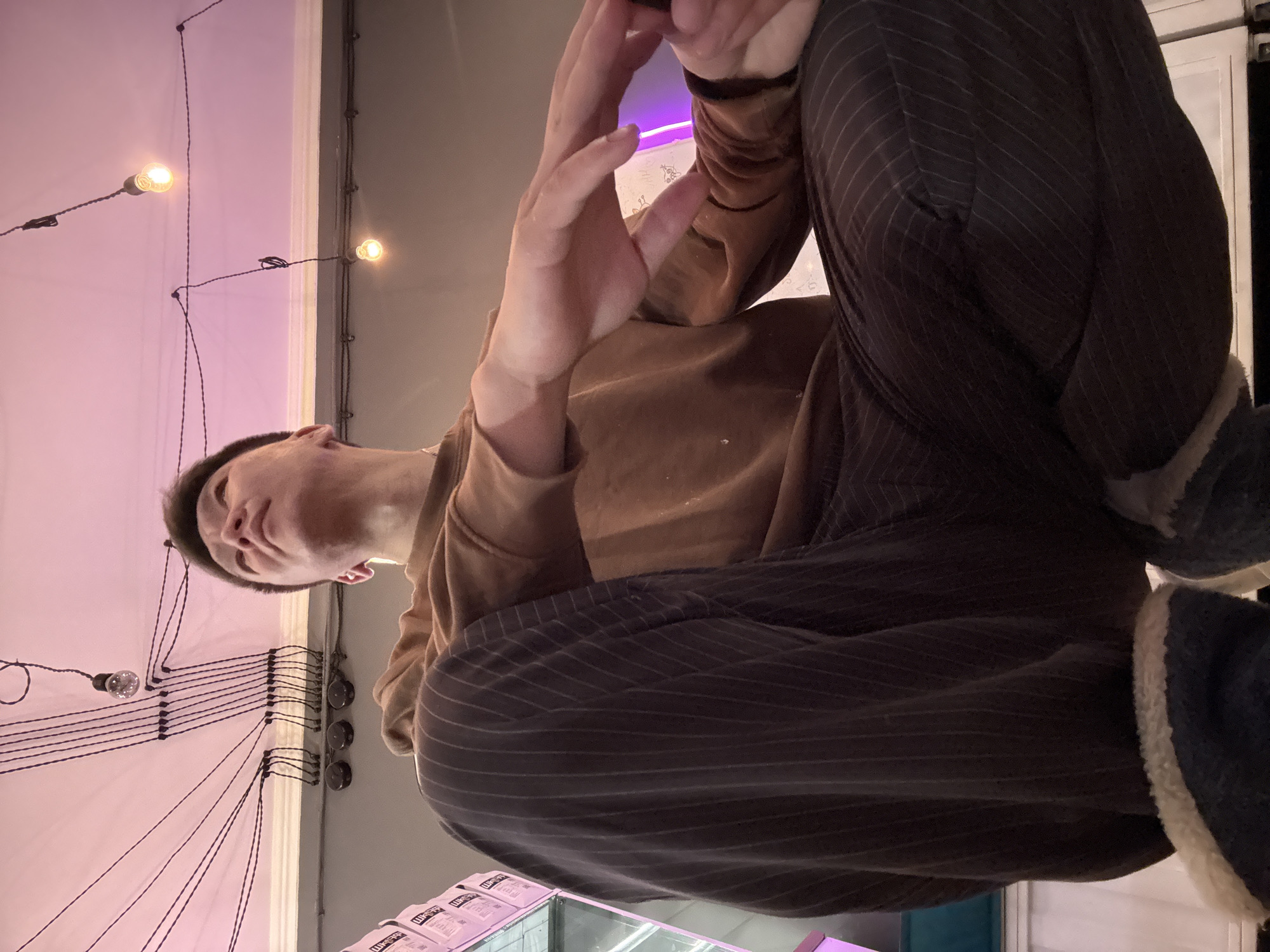 A low-angle indoor shot shows a person wearing striped pants and a brown long-sleeved shirt. The person is seated with their legs crossed. Above the person are light bulbs hanging from the ceiling and attached to wires. The background includes a wall with a purple hue. A shelf with merchandise is partially visible in the foreground. The city and location are not discernible from the image.