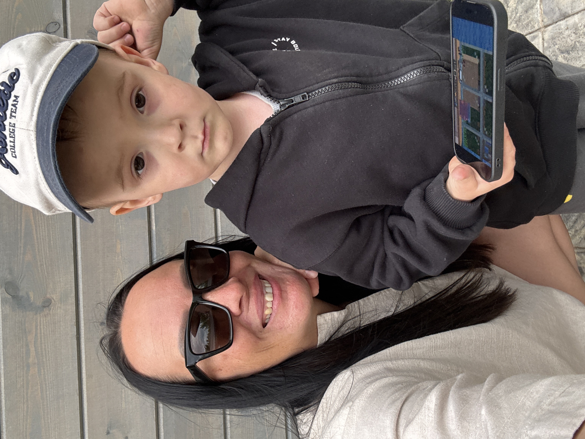 A woman with black sunglasses and black hair is smiling while holding a child with a baseball cap, wearing a black zip-up hoodie, and holding a smartphone. The background appears to be a wooden fence. The location is Chaikovsky, Russia.