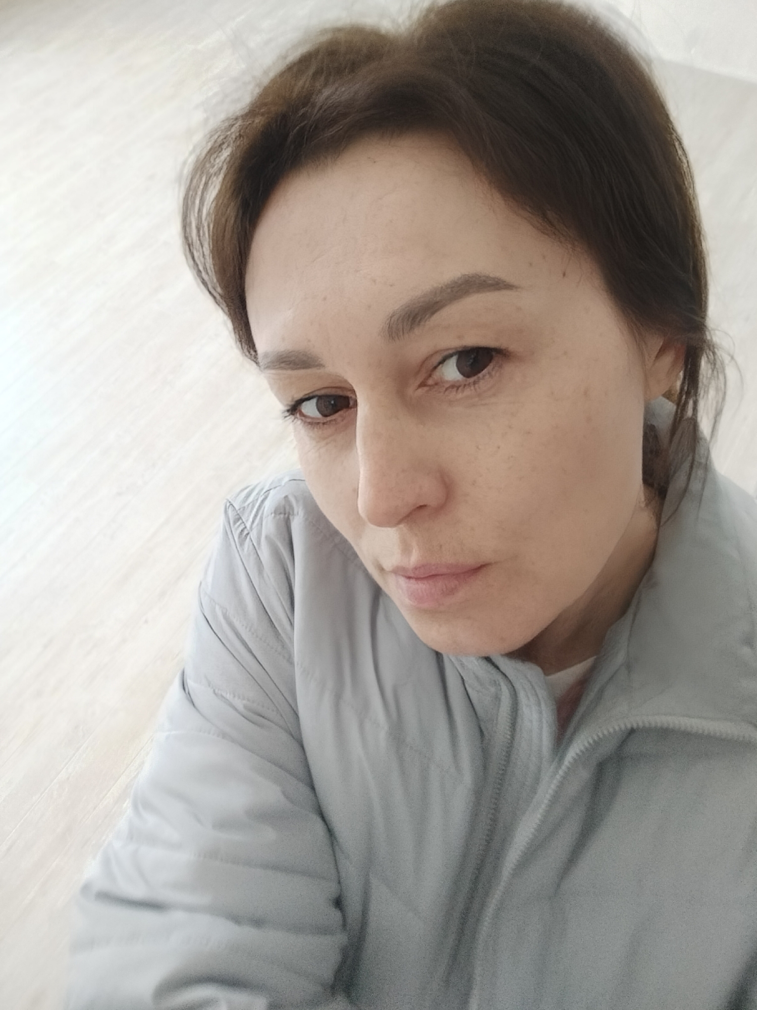 A woman with brown hair pulled back is looking down and to the left. She has brown eyes and freckles on her face. She is wearing a light gray puffer jacket with a zipper. The background appears to be a light-colored wooden floor. The image was taken in Прикамский, Russia.