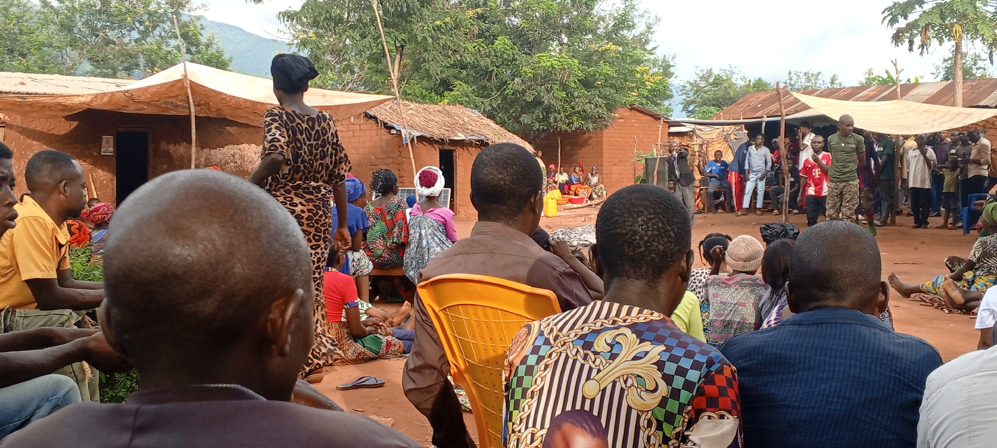Captured in Ngalula, Democratic Republic of the Congo, the image depicts a gathering of people in an outdoor setting. The scene includes buildings, likely residential, constructed of earthen materials and roofed with thatch or corrugated metal, some covered by makeshift sunshades.
Individuals of varying ages and genders are present, some seated and others standing. The seated individuals appear to be attending to a group gathered at the center, possibly listening to a speaker. The clothing worn is varied, ranging from casual attire to patterned fabrics. A limited number of chairs are visible. The environment appears to be a village or rural setting, as suggested by the vegetation and construction styles.