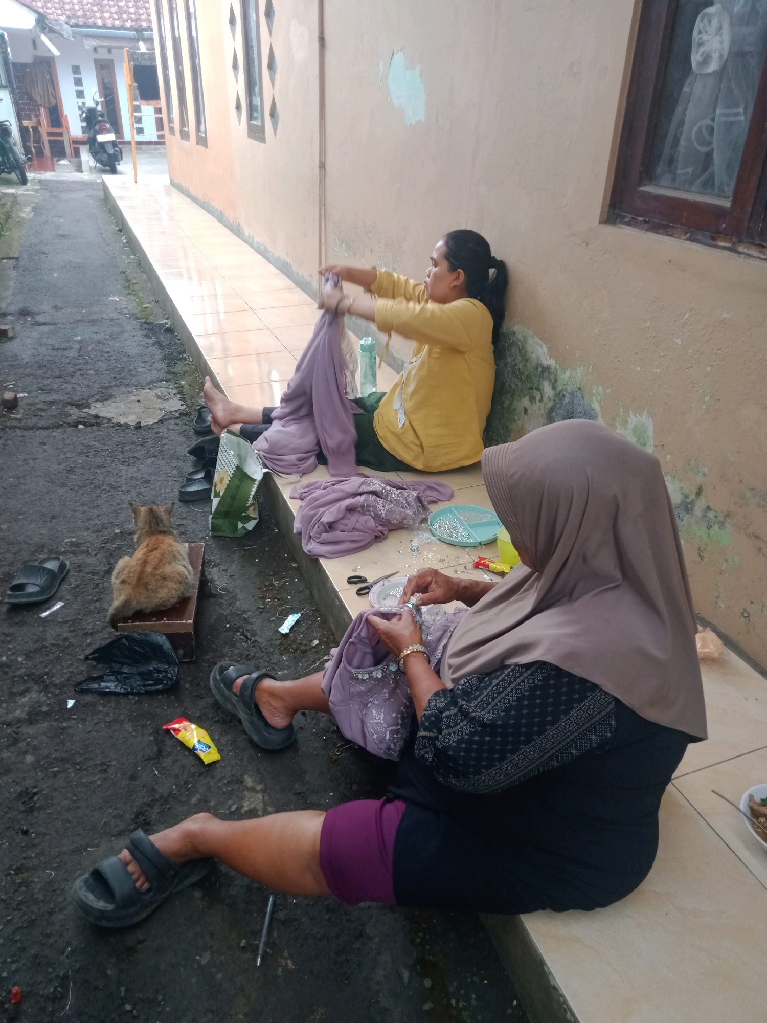 In Cintaraja, Indonesia, two women are engaged in sewing activities near a textured, light brown wall. The woman on the right, wearing a light brown hijab and a dark patterned top, is seated on a tiled ledge and is sewing intricate beadwork onto a purple fabric. In front of her are scattered beads in a light blue container, scissors, and other sewing accessories. A second woman, wearing a yellow long-sleeved shirt, is seated further back on the same ledge, holding and manipulating a piece of purple fabric. A ginger cat is sitting on a small wooden box on the ground to the left of the women. The ground area is a mix of asphalt and dirt, with scattered debris including a green and white bag, black plastic bags, and a yellow snack wrapper. A motorcycle is partially visible in the background, parked near a building with a tiled roof. The overall environment appears to be an outdoor alleyway or passage between buildings.