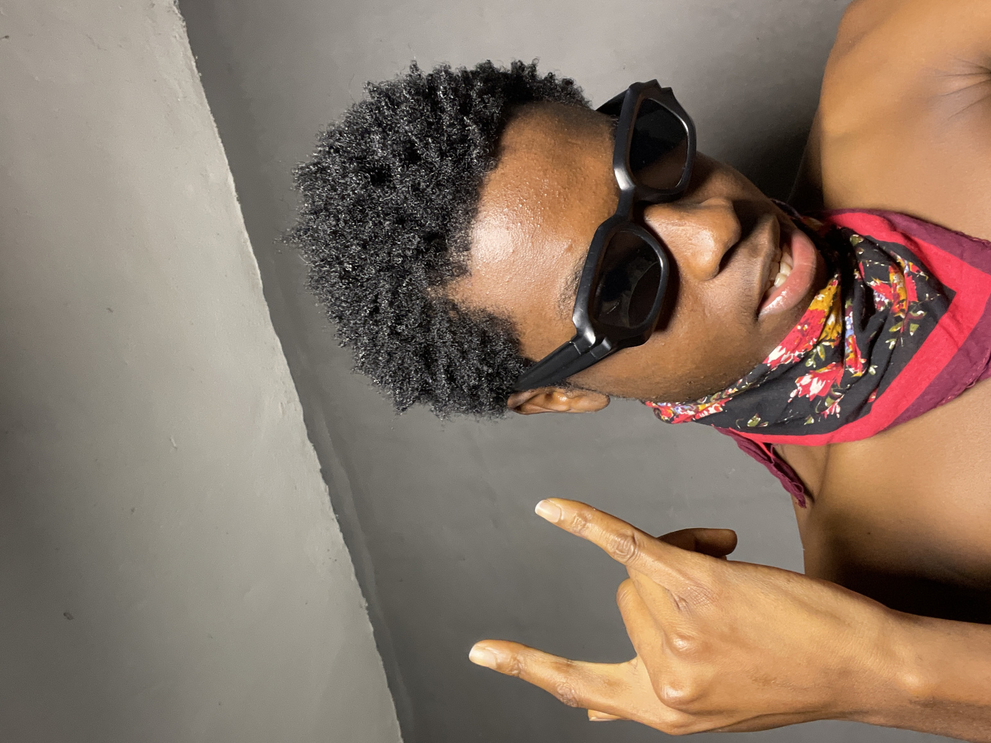 The media file features a close-up, sideways portrait of a young Black man with dark, curly hair, wearing black sunglasses and a bandana with a floral pattern and red trim. He is looking towards the camera with a slight smile, revealing his teeth. His right hand is raised, making a rock-and-roll hand gesture, with his thumb extended and index and pinky fingers raised. The man is shirtless, and his skin appears to be sweating. The background is a plain, dark gray wall, with a lighter gray, possibly concrete, surface on the left. The lighting suggests an indoor setting with artificial light. The overall mood is confident and casual. The location is identified as Nkpor, Nigeria.