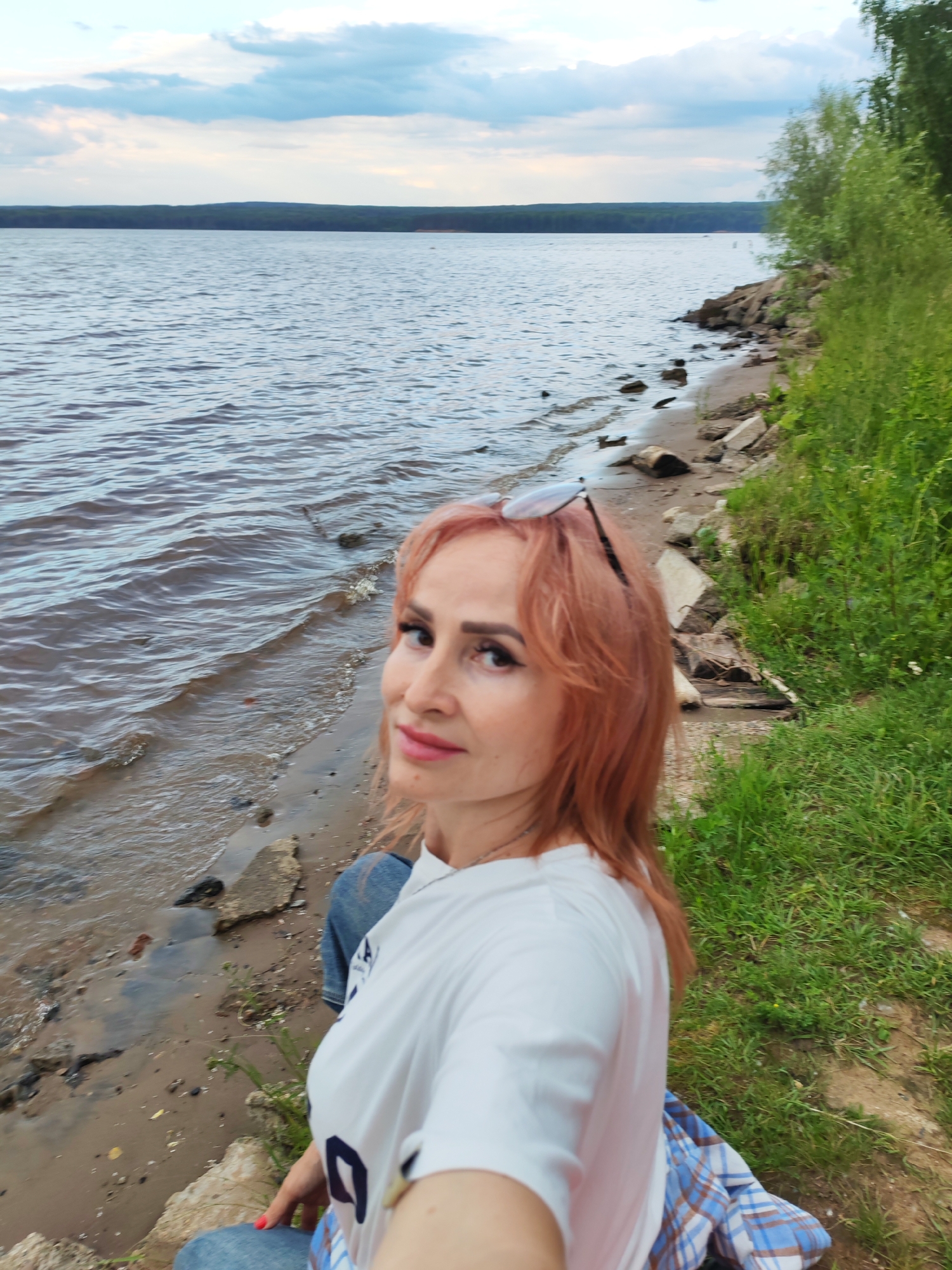A woman with pink hair, wearing a white t-shirt and blue jeans, is captured in a selfie taken from a low angle. She is seated on the shore of a body of water, possibly a reservoir, with a line of trees and a hazy horizon in the background. The water's surface is rippled, and the shore is a mixture of sand, small rocks, and some scattered debris like driftwood. Lush green grass and foliage grow along the bank to the right of the frame, suggesting a natural, outdoor setting. The sky above is partly cloudy with a soft, diffused light, indicating it could be late afternoon or early evening. The overall impression is one of a casual, relaxed moment by the water. The location is identified as Chaikovsky, Russia.