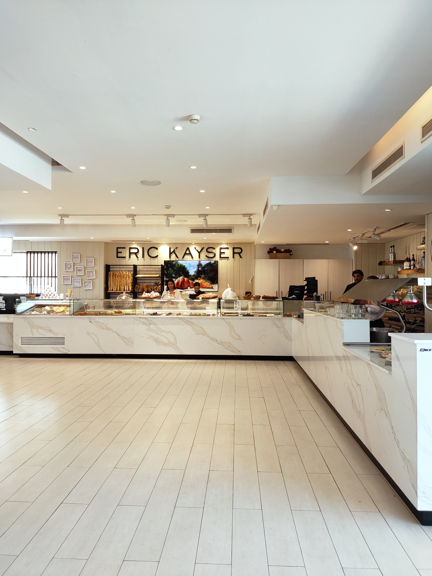 The image depicts the interior of a bakery, branded as "ERIC KAYSER," located in the city of Tunis, Tunisia. The bakery features a modern and clean aesthetic with white marble countertops and light-colored tiled flooring. A long display counter showcases a variety of baked goods, including pastries and bread. Behind the counter, two female employees are visible, one attending to a display and the other interacting with a customer. The wall behind the counter features the bakery's logo and a large screen displaying an advertisement for croissants and other baked items. In the background, shelves are stocked with bread loaves, and additional storage cabinets are present. A male employee stands near a coffee machine and other service equipment on the right side of the counter. The ceiling is white and equipped with recessed lighting and ventilation systems. The overall environment suggests a well-maintained retail space.