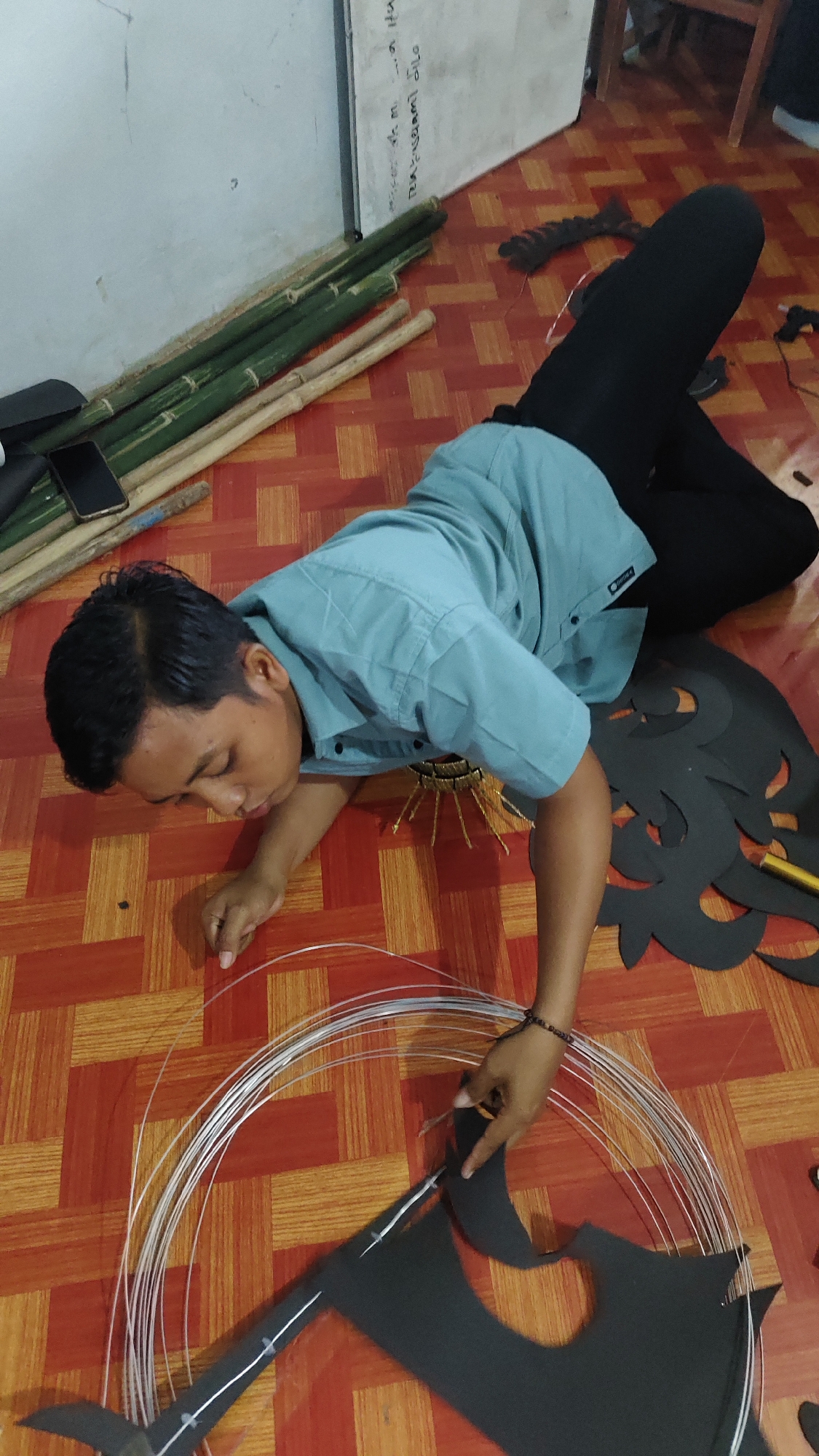 A young man in a light blue collared shirt and black pants lies on a red and orange checkered floor, looking down at his work. He is in the process of constructing a kite. In front of him are several black, intricately cut pieces of material, resembling wings or decorative elements of a kite. A bundle of thin, white wire is coiled and spread out, being manipulated by his hands. To his left, a stack of long, green bamboo poles leans against a white wall. A black mobile phone rests on top of the bamboo. The environment appears to be indoors, possibly a workshop or a room in a residential setting within Banyubunih, Indonesia, suggested by the presence of bamboo and the typical flooring pattern. A wooden chair is visible in the background.